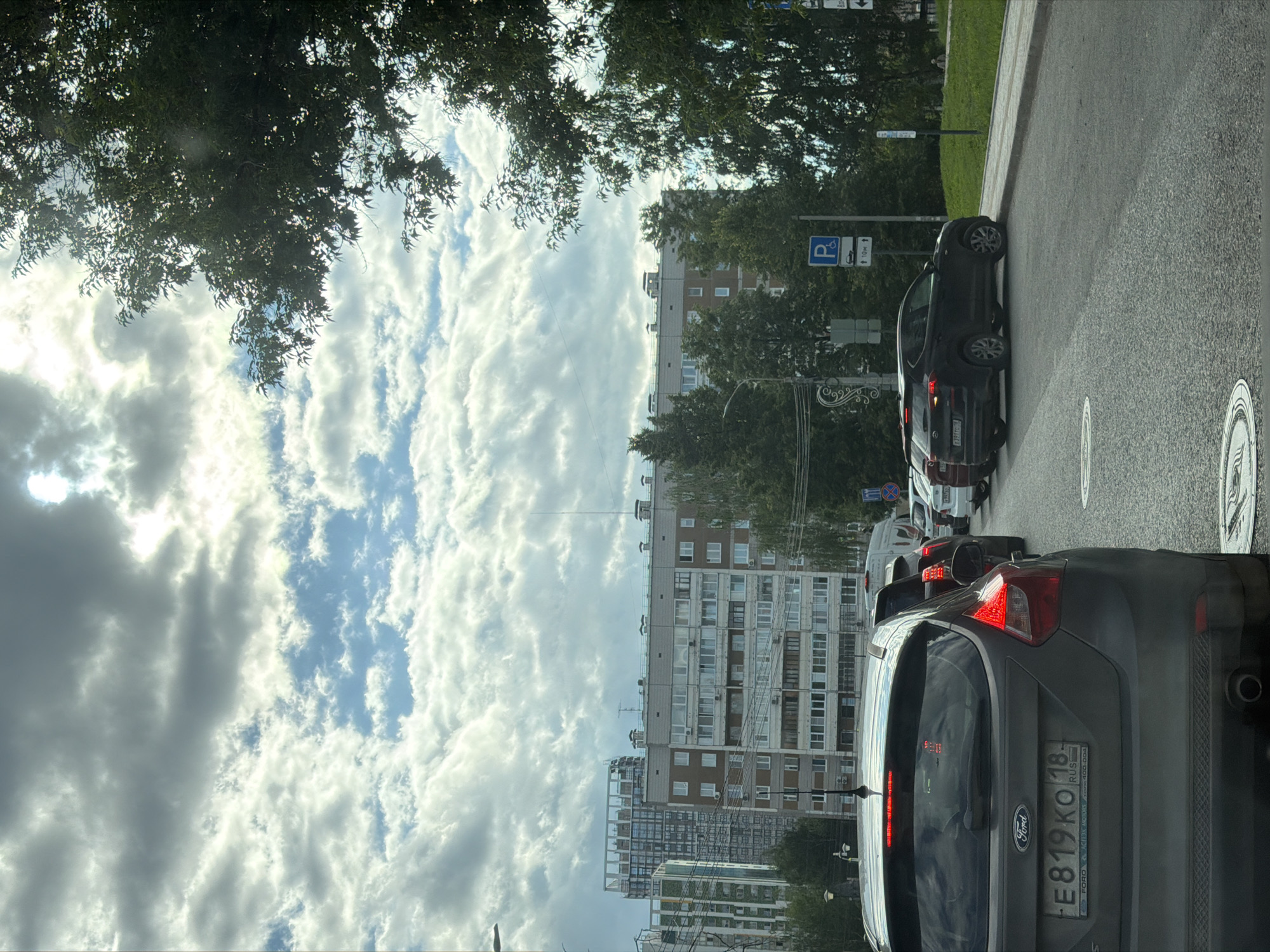 The image captures a street scene in Izhevsk, Russia, likely during daytime with overcast skies and scattered sunlight. In the foreground, the rear of a dark gray Ford Focus is visible, displaying a Russian license plate that reads "E819KO 18 RUS". The car is part of a traffic jam, with other vehicles lined up behind it, including a black Hyundai Tucson parked on the right side near a curb, and further back, several other cars and a white van.
The urban environment includes multi-story apartment buildings in the background, characteristic of Russian architecture, with many windows. Tall trees, some with dense foliage, line the right side of the street, partially obscuring the buildings. A parking sign with a wheelchair symbol and arrows indicating direction and distance is visible on a pole. The road surface is asphalt, with a circular manhole cover prominent in the lower foreground. The overall impression is a typical urban traffic situation under a partly cloudy sky.