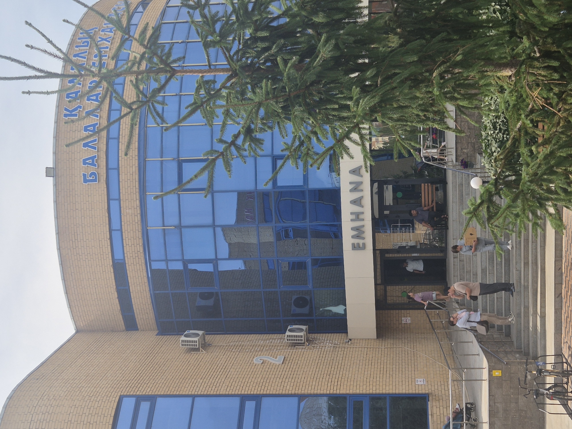 The image shows the exterior of a modern building, likely a medical facility or a public institution, with a distinctive curved facade. The building is constructed with light-colored brick and features large, reflective blue windows. A prominent sign above the entrance reads "EMHANA," and further up on the curved facade, there is lettering in blue that appears to be in a Cyrillic-based script, possibly "ҚАЗАҚ ТІЛІ БАЛАЛАРҒА АРНАЛҒАН" which translates to "KAZAKH LANGUAGE FOR CHILDREN." The location is identified as Kokshetau, Kazakhstan.
In the foreground, several people are visible on a set of wide stairs leading up to the entrance. Some are standing and talking, while others are walking or carrying items. On the left side of the stairs, a baby in a stroller is positioned on a ramp. To the right of the stairs, a bench is placed near the entrance, with a person sitting on it. A large evergreen tree partially obscures the view of the building on the right side of the image. On the left wall of the building, two air conditioning units are mounted, and a large numeral "2" is displayed. Bicycles are parked in the lower left corner of the image. The sky is overcast.