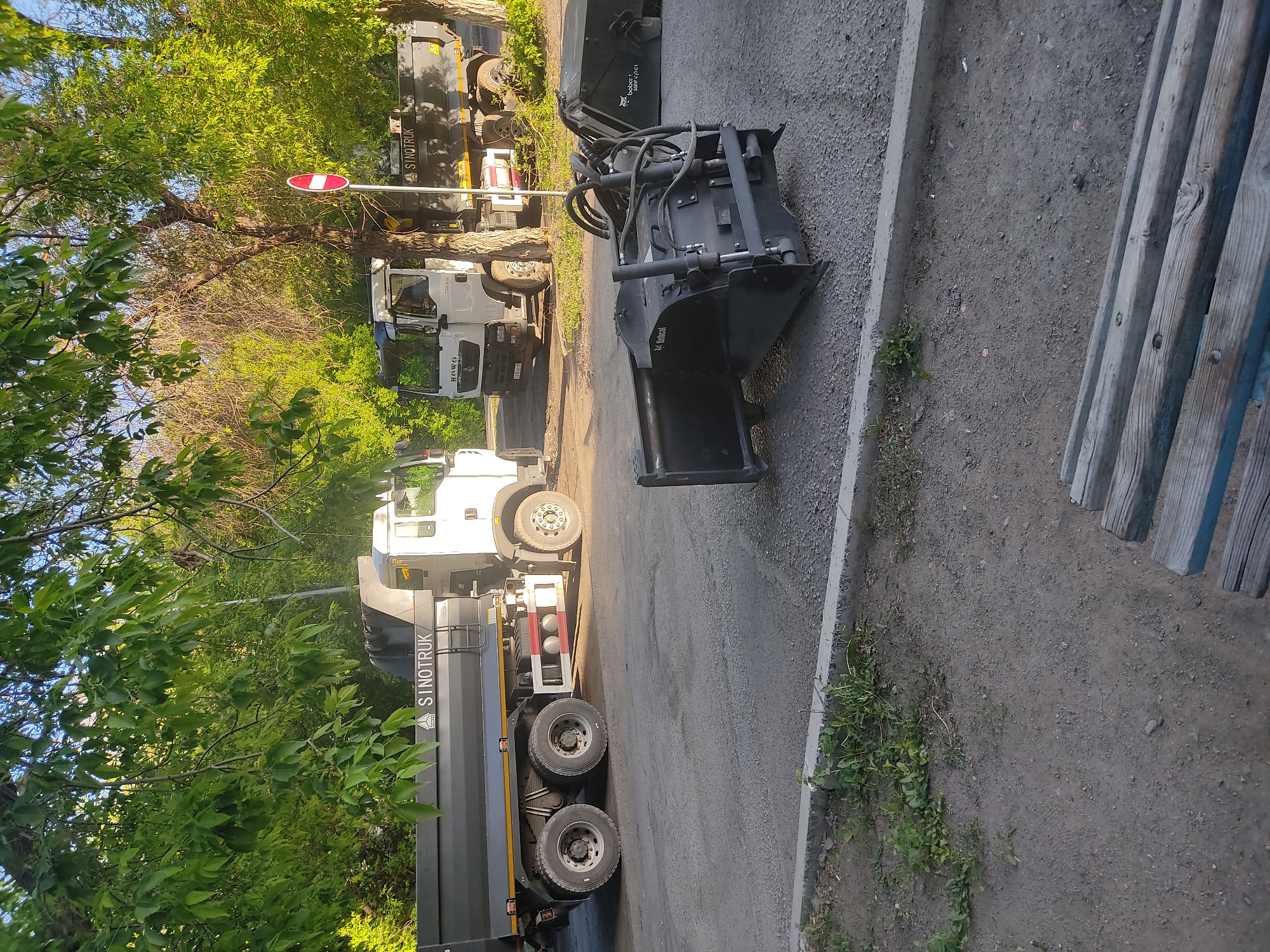 The scene depicts a street in an unknown city with several construction-related vehicles. On the left side of the frame, there are three trucks of the brand "SINOTRUK" parked along the road, obscured partially by trees. An additional truck is also visible behind them. To the right is a piece of construction equipment, a black Bobcat attachment, resting on the pavement beside a curb. Next to the curb is an object of gray wood, possibly a bench or barrier. The environment includes foliage, pavement, and signs of construction or maintenance work, implying an urban setting within a city.