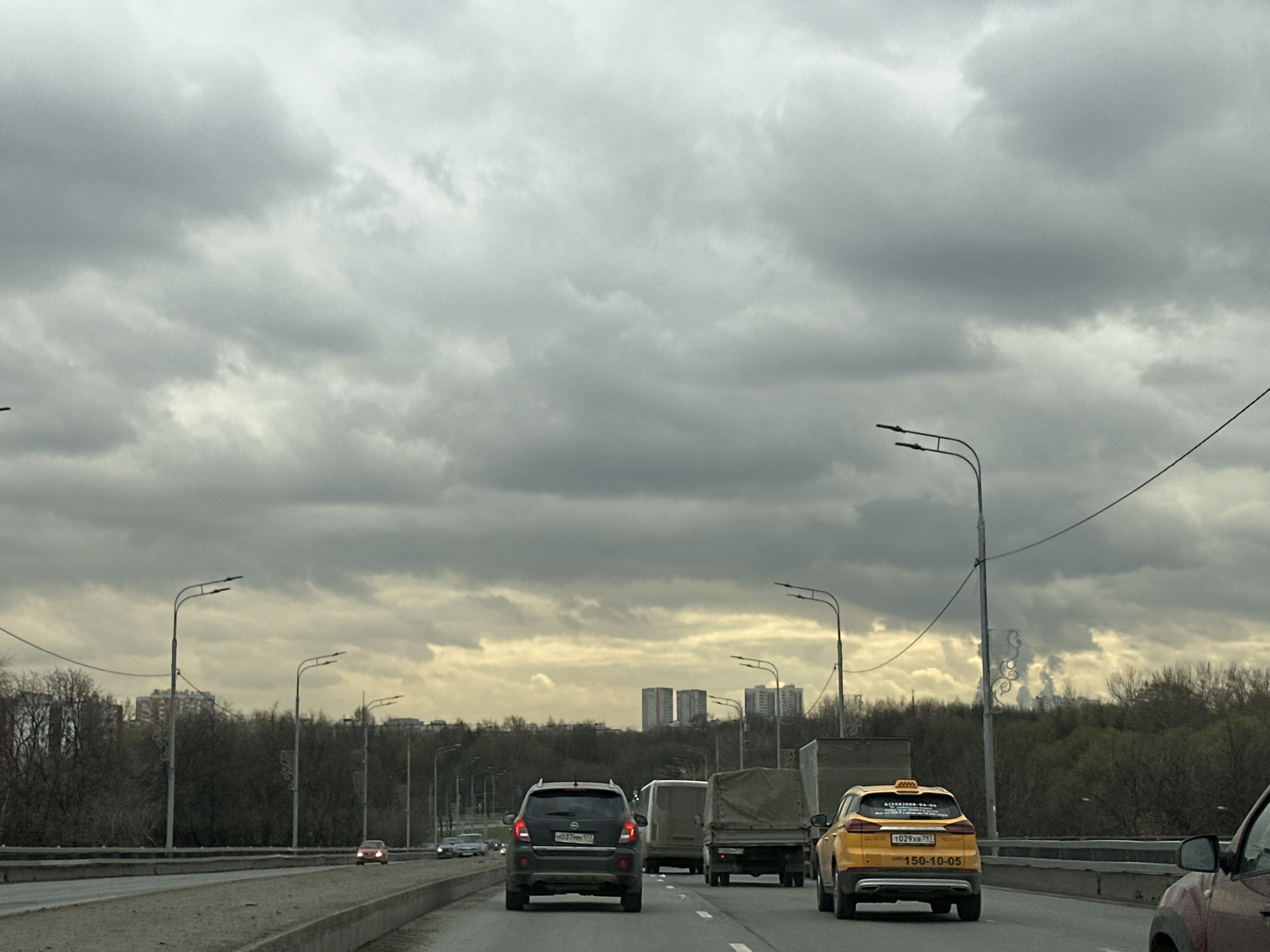 The image depicts a highway in Moscow, Russia, under a cloudy sky. Several vehicles are present, including a dark gray car, a white van, a covered truck, and a yellow taxi with the license plate "T029XB 797" and "150-10-05". The highway is flanked by trees and streetlights. In the distance, there are buildings and what appears to be smoke or steam rising.
