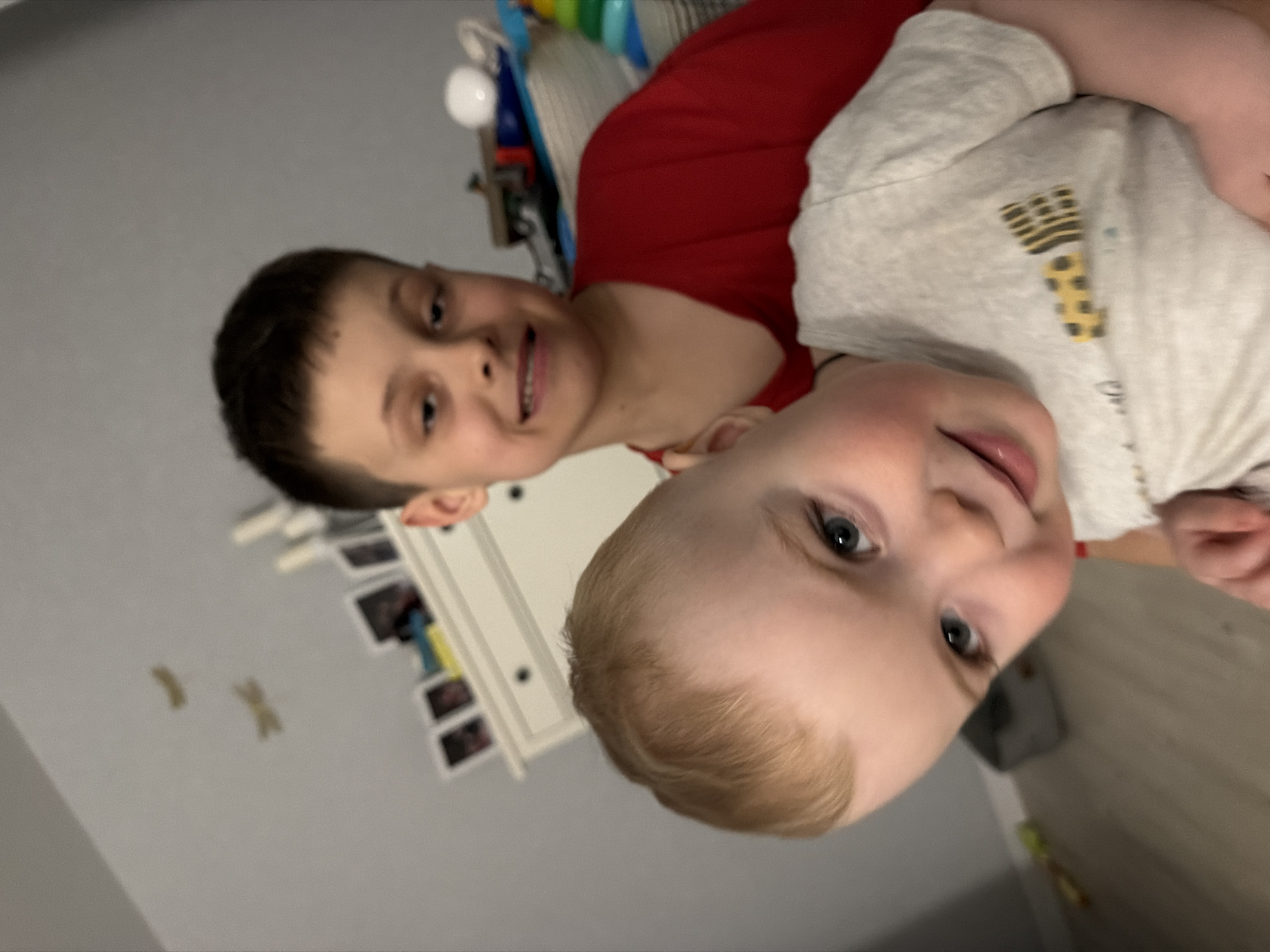 A portrait depicts two individuals, one older than the other. The older child is wearing a red shirt. The younger child is wearing a light-gray shirt. The photo appears to be taken indoors, in front of a wall with framed pictures. There is no verifiable information suggesting this image was taken in Votkinsk, Russia.