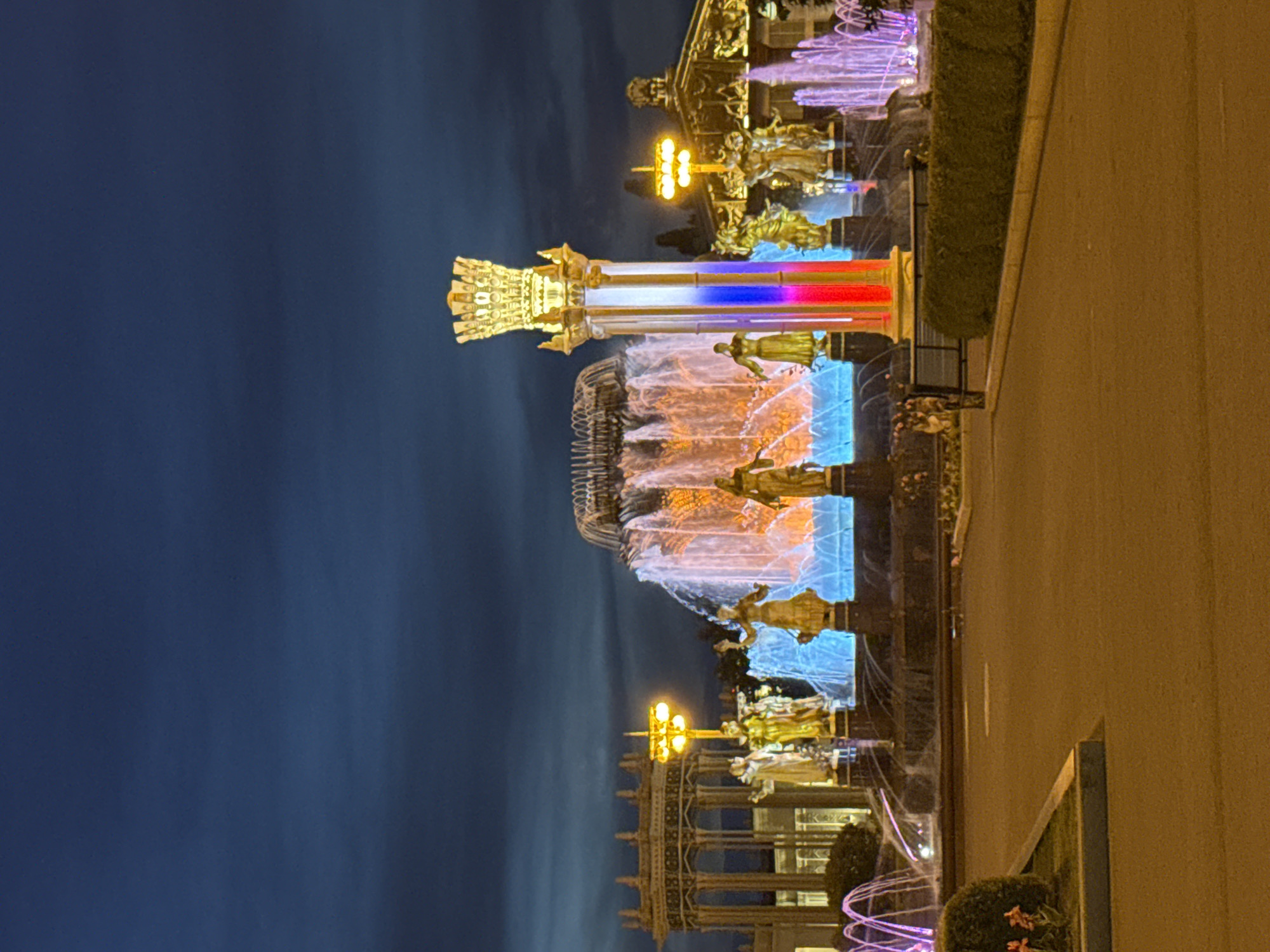 A wide-angle, low-light photograph captures the Friendship of Peoples Fountain in Moscow, Russia, at dusk. The fountain is illuminated, with cascading water glowing in shades of blue, orange, and pink. Gilded statues of people are visible around the fountain's base and on a tall, ornate column in the foreground. This central column is decorated with vertical stripes of red, white, and blue, representing the Russian flag. The sky above is a deep, dark blue, with some clouds visible. In the background, ornate buildings with arched facades and illuminated streetlights suggest a grand architectural setting. A paved pathway and a dark green hedge with a bench are in the lower foreground.