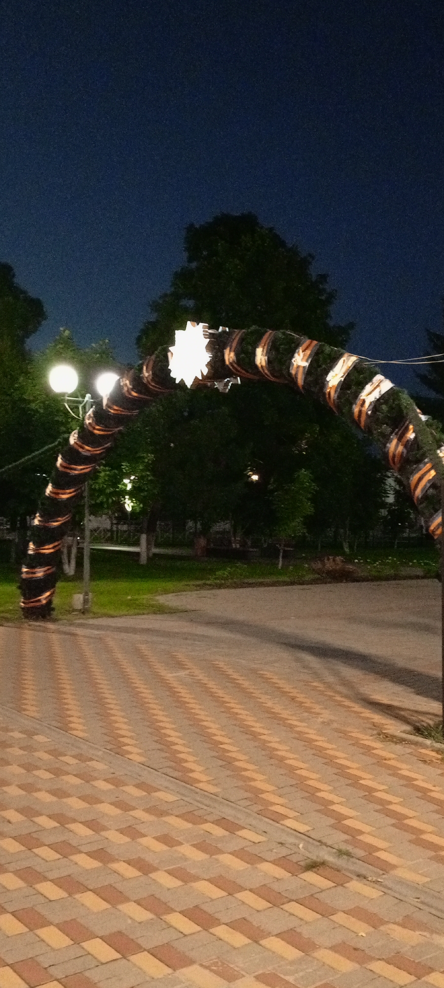 This image shows an arched structure decorated with garlands of dark green foliage and orange and black striped ribbons, possibly St. George's ribbons. A bright white star ornament is affixed to the apex of the arch. Two round streetlights with white bulbs are visible on a pole to the left of the arch, partially obscured by foliage. The arch spans across a paved walkway made of light brown and tan interlocking pavers, which then transitions into an asphalt road. Lush green trees and grass are present in the background, with a dark blue night sky overhead. The scene appears to be in a park or public space. The location is the city of Kirsanov, Russia.