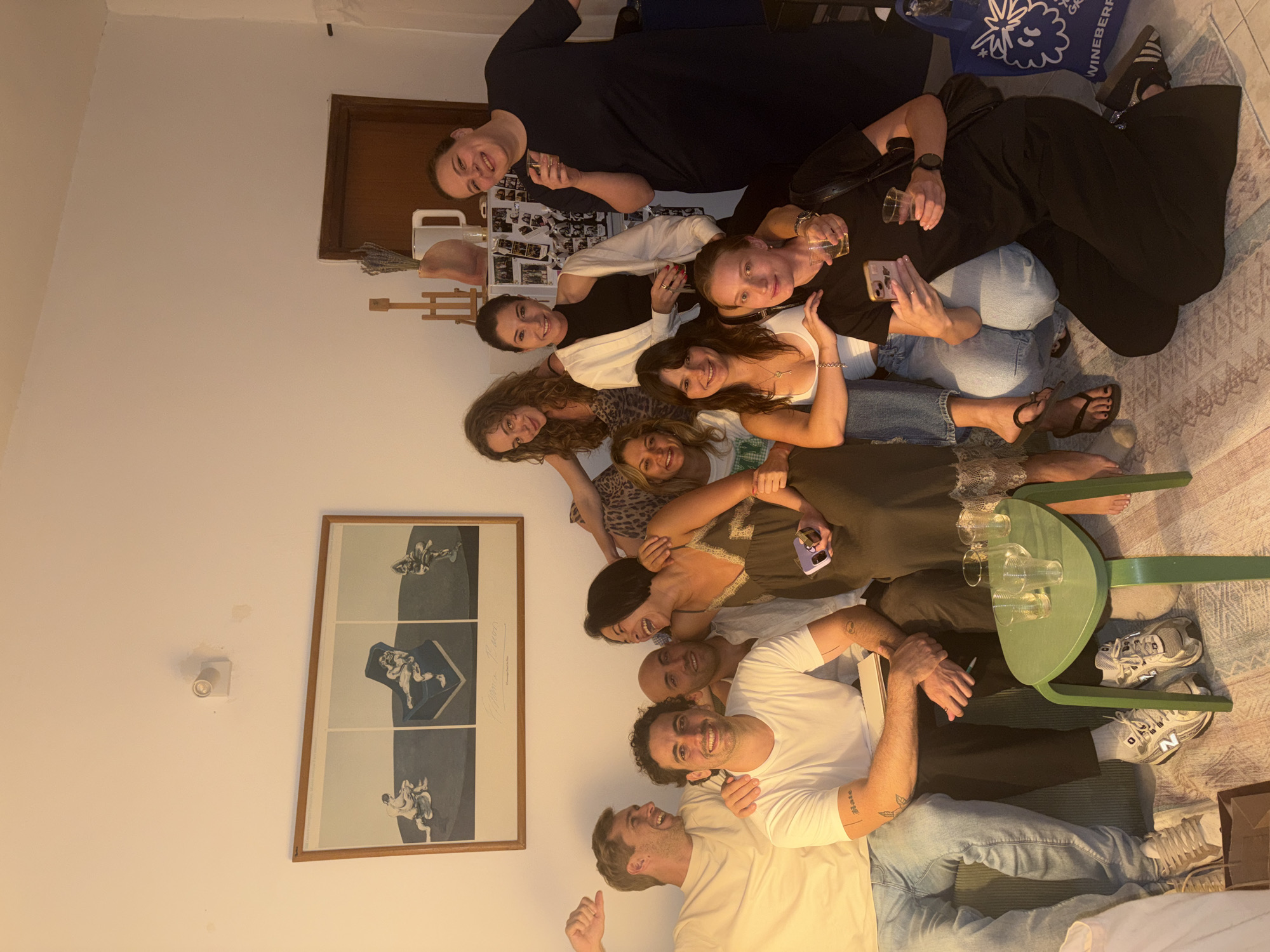 In Tel Aviv, Israel, a group of ten people, comprising six women and four men, are gathered indoors for what appears to be a casual social event. The setting is dimly lit, with the primary light source emanating from the right side of the frame, casting shadows across the room. The group is positioned around a couch and on the floor, creating a relaxed and intimate atmosphere.
Several individuals are holding drinks in their hands, suggesting a gathering with refreshments. Some people are engaged in conversation, indicated by their facial expressions and postures. One woman is captured in mid-laughter, her head tilted back, while others smile and interact with each other. A man on the left is making a gesture with his thumb up, possibly expressing approval or enthusiasm.
The environment includes a couch, a small green table with glasses on it, and a framed artwork hanging on the wall. The artwork depicts three stylized figures in a monochromatic palette. In the background, a doorway and what appears to be a refrigerator adorned with photographs are visible. The floor is covered by a patterned rug.
The people are dressed in casual attire, ranging from t-shirts and jeans to dresses. Their poses and expressions convey a sense of camaraderie and enjoyment. The overall impression is of a convivial social gathering among friends in a home setting.