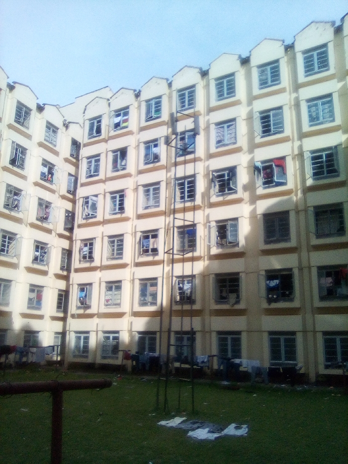 This image presents a multi-story residential building located in Bondo, Kenya, viewed from a ground-level grassy courtyard. The building is constructed of a light yellow/cream-colored material with brown vertical structural accents and exhibits a curved or angled architectural footprint. It rises at least six stories, featuring a distinctive roofline with repeating stepped parapet elements above the top floor windows. Numerous multi-pane casement windows are visible across all floors, with many partially open. Laundry items, primarily clothing, are visible hanging from or protruding out of several open windows on various levels. A tall, dark metal structure, resembling a ladder or scaffolding, stands vertically in the central courtyard area, extending upwards multiple floors. The foreground consists of a well-maintained green lawn. Several pieces of white and dark clothing are laid out flat on the grass to dry. Additional laundry is discernible hanging on lines or railings at the base of the building, adjacent to the grass. A partial brown metal bar is visible in the lower-left quadrant. The sky is clear and light blue. No human figures are present in the scene.