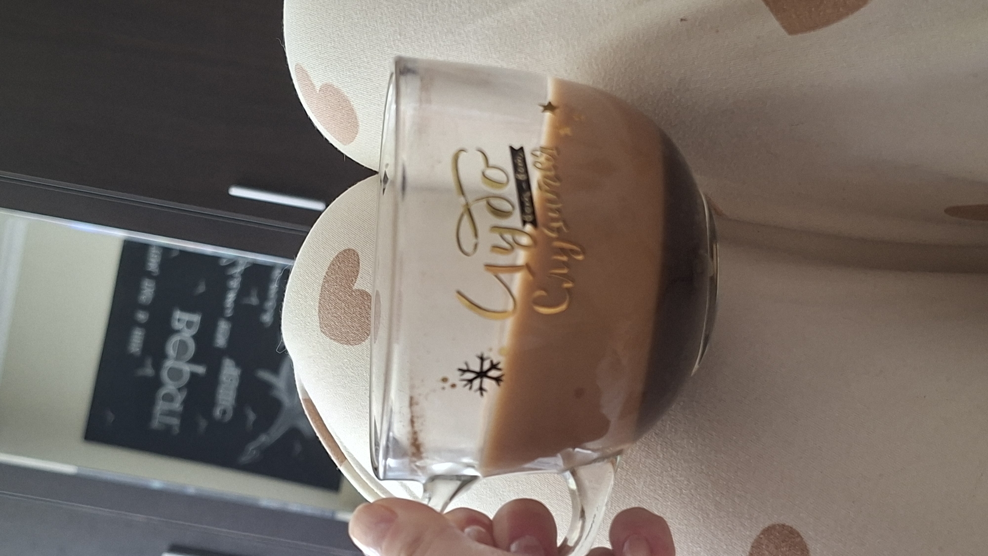 A clear glass mug filled with a layered beverage, likely coffee or hot chocolate, is held by a person's left hand. The mug is positioned on a light-colored, textured surface, possibly a piece of clothing with a subtle heart pattern. The beverage has a dark bottom layer and a lighter, frothy top layer. Gold lettering is visible on the mug; the top line reads "Чудо" (Chudo), meaning "miracle," and the bottom line reads "Случится" (Sluchitsya), meaning "will happen." A small snowflake graphic is also present on the mug. In the background, a reflection in a mirror shows an unfocused image of a dark board with white lettering. The overall setting suggests a casual, indoor environment. The specific location in the city of Myadzyel, Belarus cannot be determined from the visual information provided.