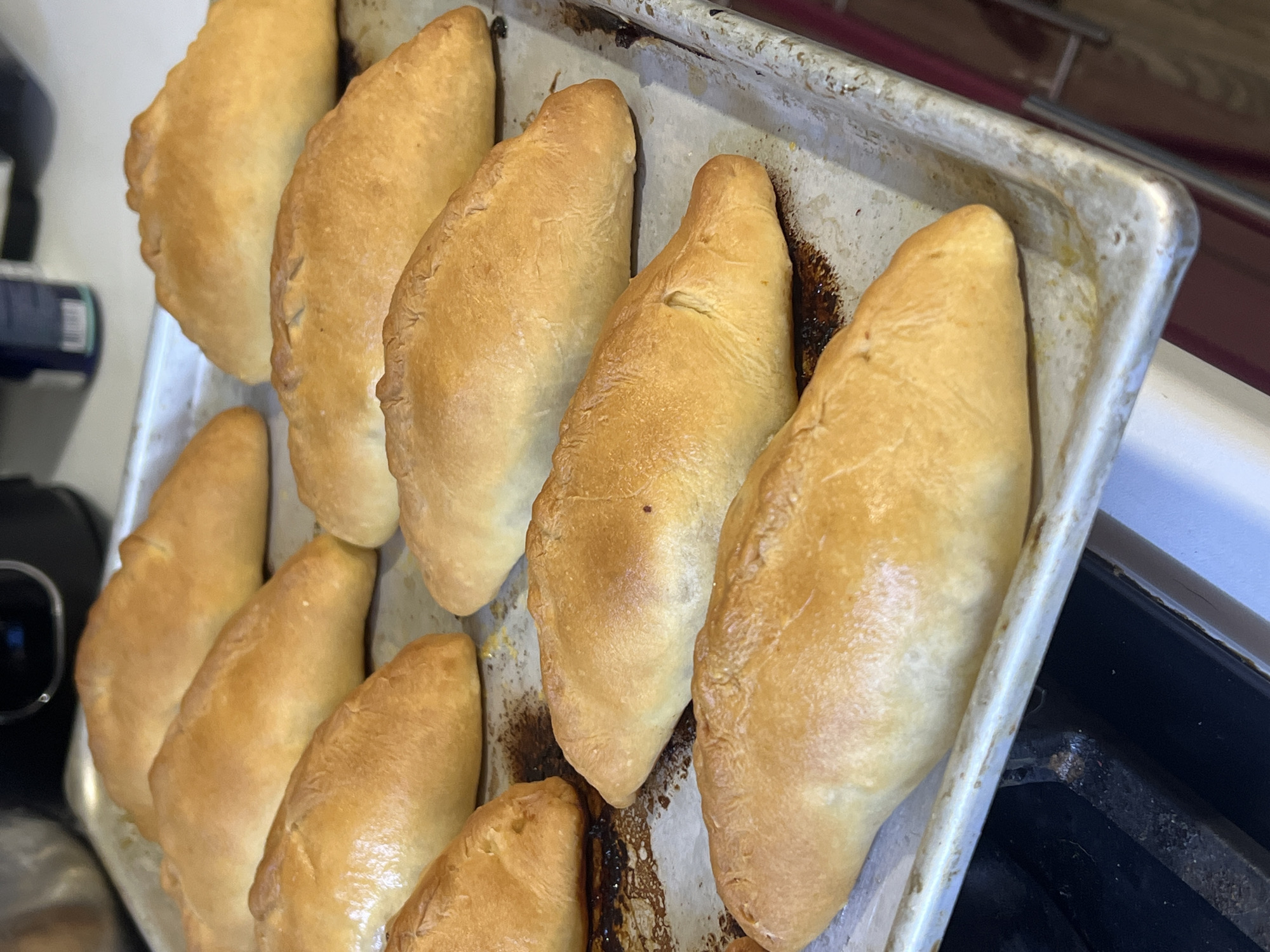A metal baking sheet containing approximately ten golden-brown, oval-shaped baked goods, possibly pirozhki or similar pastries, is positioned on a stovetop. The baking sheet shows signs of previous use, with darkened areas indicating baked-on residue. The baked goods are arranged in rows, with some slight variation in size and shape. The stovetop appears to be part of a white oven unit, and the background includes partially visible kitchen appliances and countertops. The location in the city of Noviy, Russia, is not evident from the visual information provided.