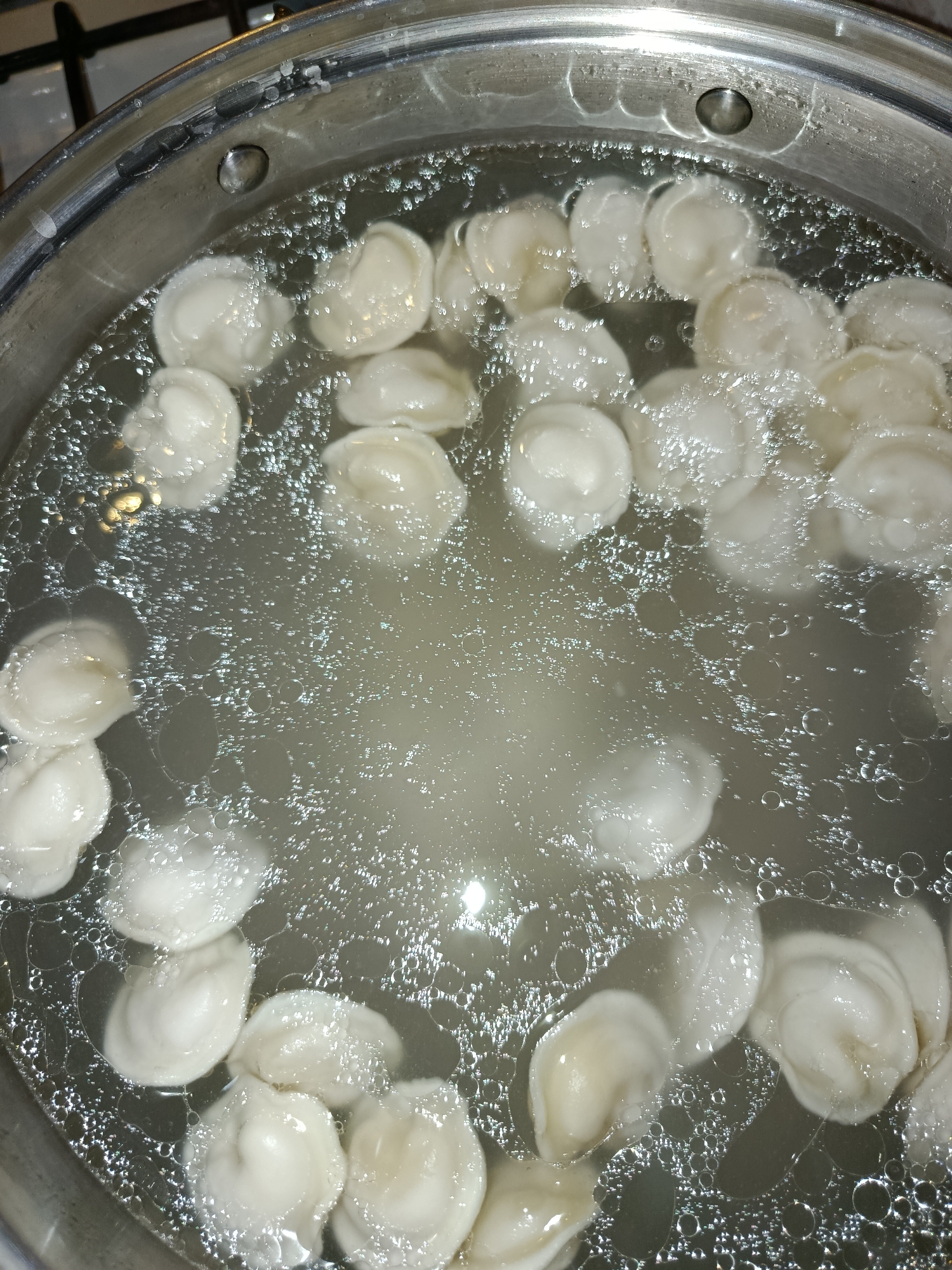 This is a close-up, top-down shot of pelmeni (Russian dumplings) boiling in a silver pot. The pot is filled with clear, bubbling water, and a multitude of pelmeni are submerged and floating. The pelmeni are small, white, and crescent-shaped, with visible crimped edges. Some are clustered together, while others are more spread out. The water's surface is agitated, with many small bubbles rising to the top, indicating an active boil. The lighting is somewhat dim, with reflections visible on the water's surface and the pot's rim. The scene appears to be set in a kitchen, likely during meal preparation. The overall impression is one of a home-cooked meal in progress. No people, text, or specific time of day cues are visible. The location context of Tashkent, Uzbekistan, suggests this dish is being prepared in a Central Asian home.