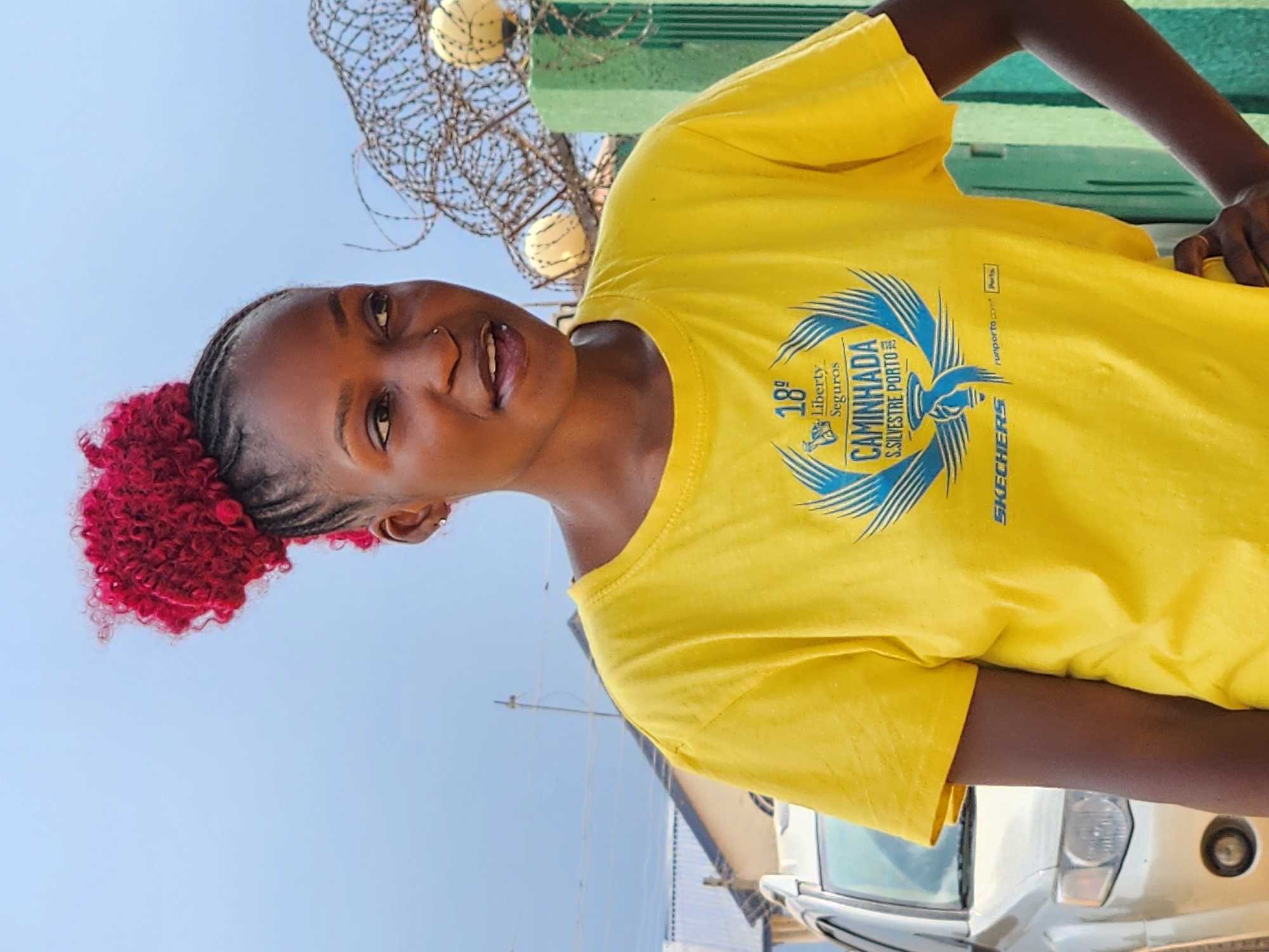 A young woman with dark skin and bright red, curly hair styled in a bun stands outdoors in what appears to be the city of Sabon Tasha, Nigeria. She is wearing a bright yellow t-shirt with a blue graphic design. The graphic features a torch with wings and text that includes "18° Liberty Seguros CAMINHADA S.SILVESTRE PORTO" and "SKECHERS". Her left hand is resting on her hip, and her right hand is not fully visible. In the background, a light-colored SUV is parked, and a green fence with barbed wire and spherical objects is visible. The sky is a pale blue, and some buildings and utility poles can be seen in the distance.