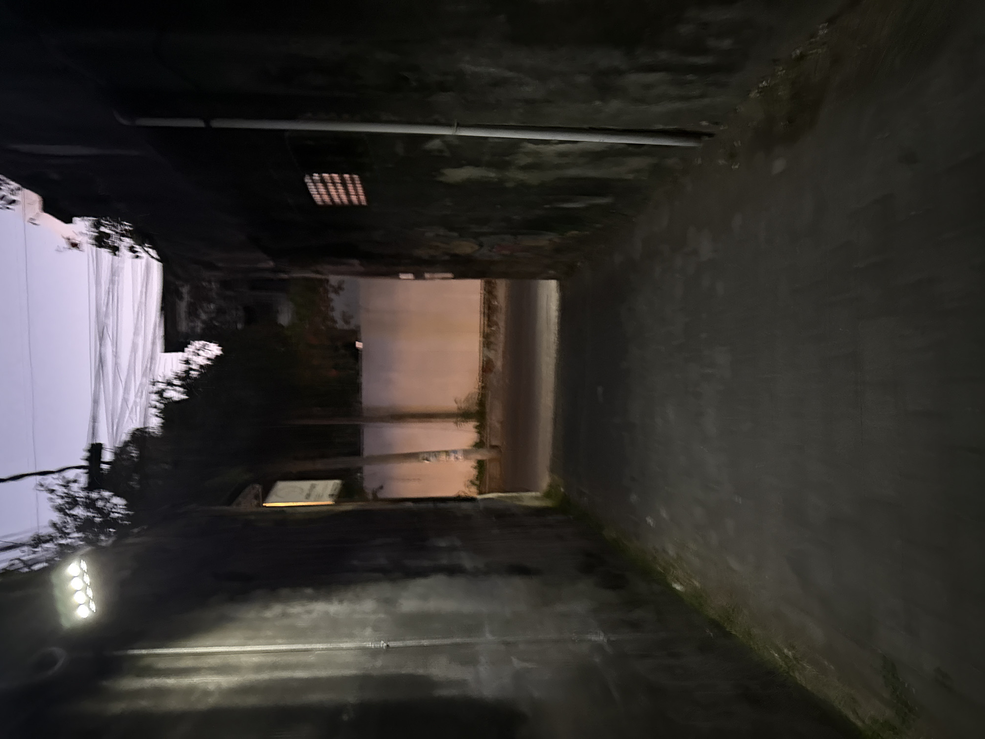 This is a low-light, outdoor image of a narrow alleyway in Denpasar, Indonesia. The alley is flanked by dark, textured walls on both sides. On the left wall, a modern, rectangular LED light fixture illuminates a portion of the alley. The right wall features a small, grated window with light emanating from within. A white pipe runs vertically down the right wall. The ground is paved with a dark asphalt material. At the end of the alley, a street is visible with a light-colored wall in the background, possibly part of a building or fence. Trees and electrical wires are silhouetted against a dusky purple sky, suggesting the image was taken at twilight. The overall atmosphere is dimly lit and somewhat enclosed.
