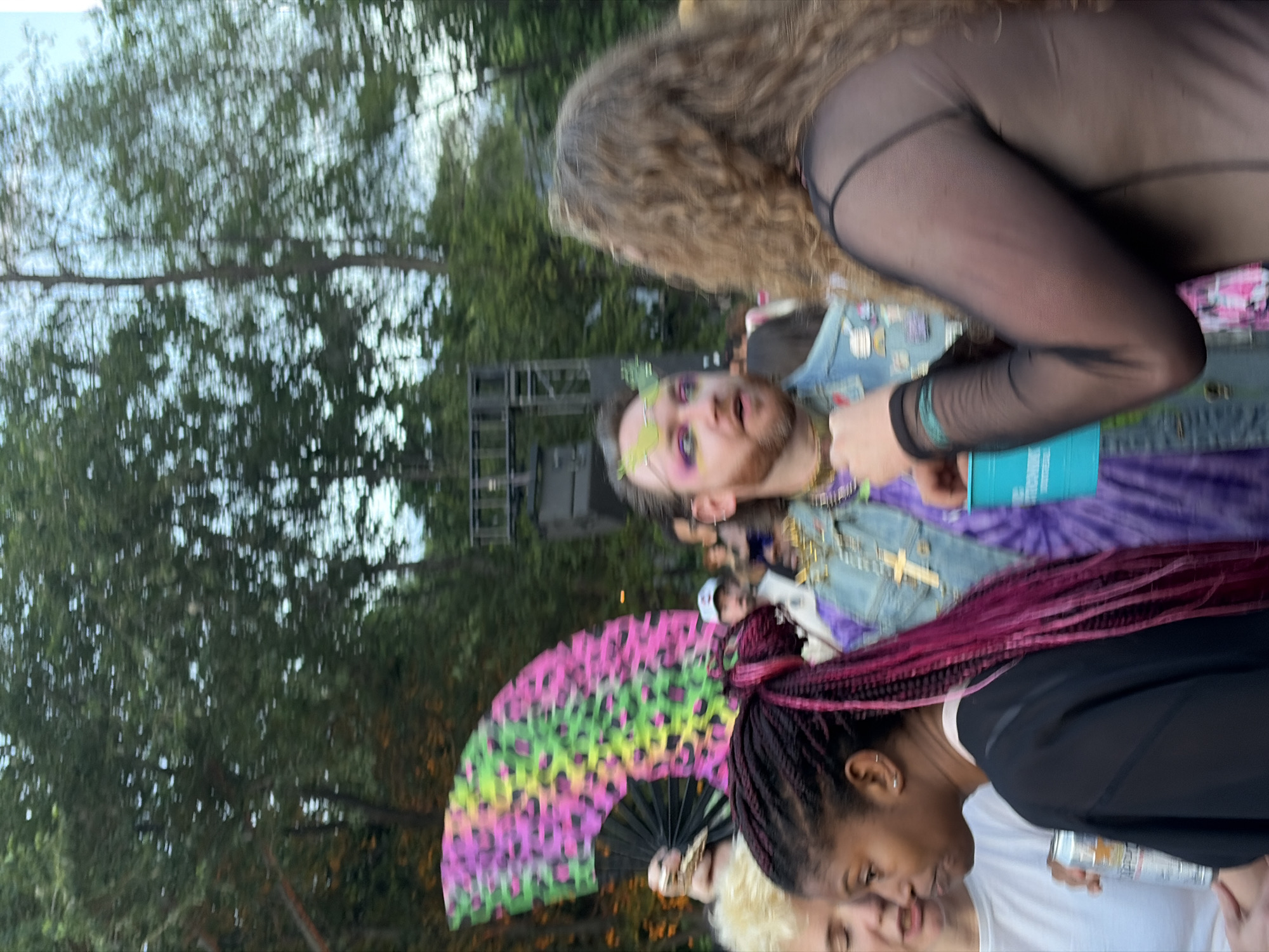 A group of people are gathered outdoors, possibly at a festival or event, with trees and a stage-like structure in the background. In the foreground, a person with dark braided hair and pink streaks holds a silver can. To their left, another person's face is partially visible. In the center, a person with eye makeup and facial hair wears a denim jacket adorned with pins. To the right, a person with curly hair is seen from the back, wearing a sheer black top. A large, colorful fan with a leopard print pattern is held by someone in the background. The overall impression is of a vibrant and festive atmosphere. The image was taken in Montreal, Canada.