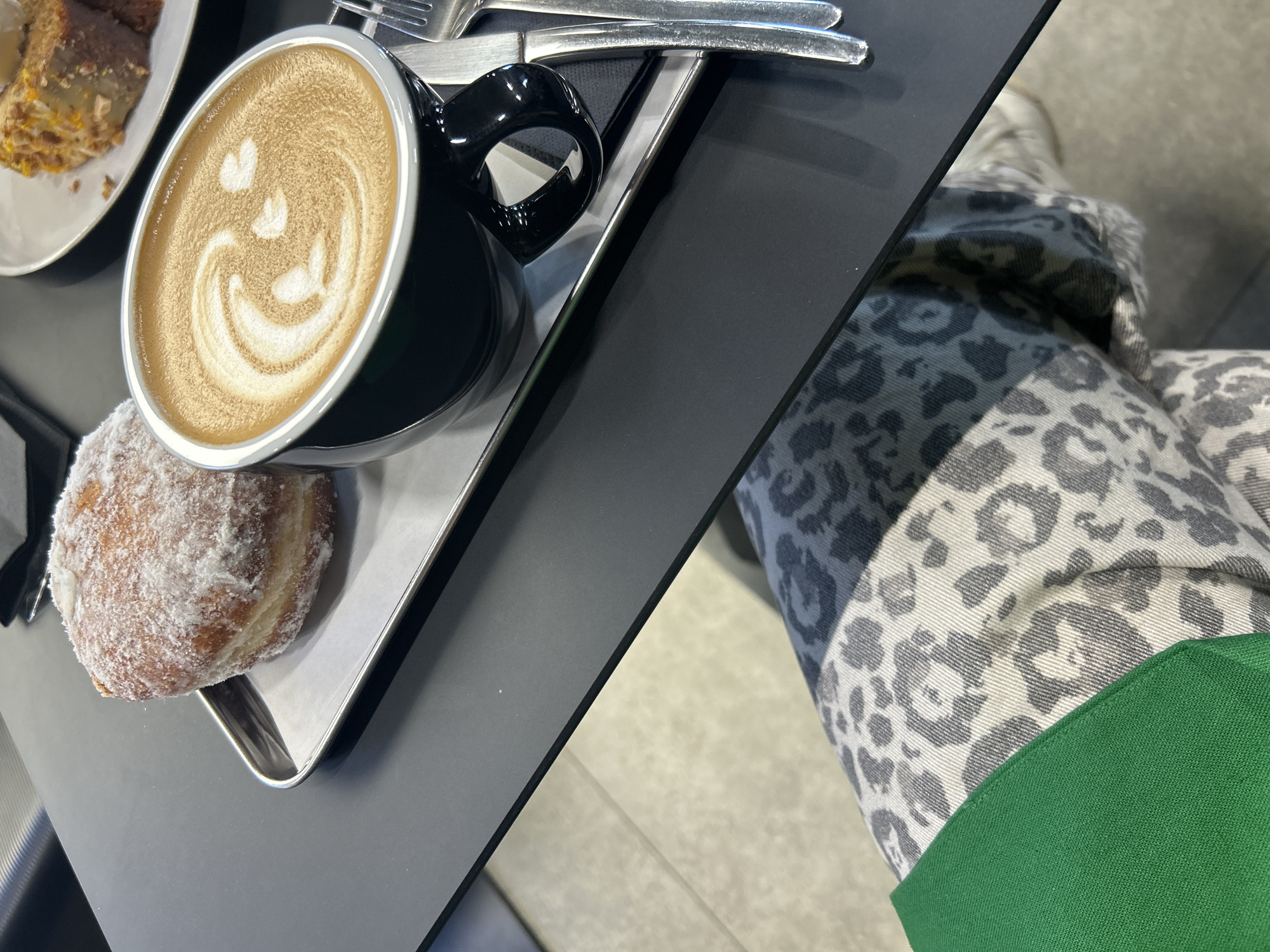 The image shows a black table surface in an indoor setting. On the left side of the table, there's a silver tray holding a cup of coffee with latte art resembling a smiling face, a powdered donut, a plate with a piece of cake, and silverware. The right side of the table surface is plain black. A person wearing gray and white leopard-print pants with a green garment visible appears to be sitting beside the table. The setting resembles a cafe. There is no information in the image to determine the location in the city of Izhevsk, Russia.