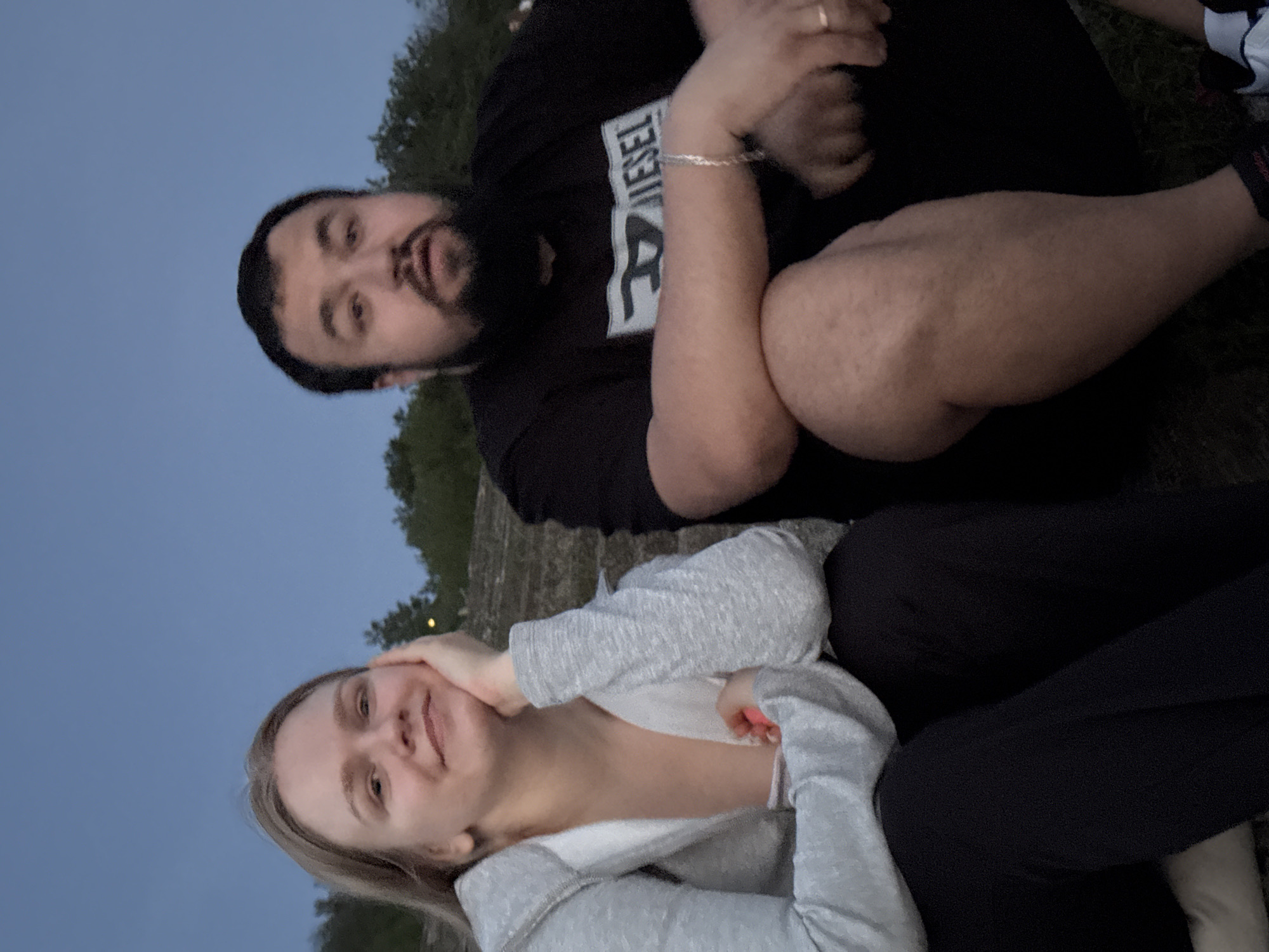 Two individuals are positioned outdoors. A female with fair skin and blonde hair is on the lower portion of the frame, wearing a light-colored hooded sweatshirt and dark pants. She rests her hand against her cheek. A male with a beard and dark hair is on the upper portion of the frame wearing a dark-colored t-shirt. The environment appears to be an outdoor location, possibly a park or open space, as suggested by the presence of trees in the background. The sky is visible, with a color suggesting either dusk or dawn. The exact location within the city of Unknown, Unknown is not determinable based on the image.
