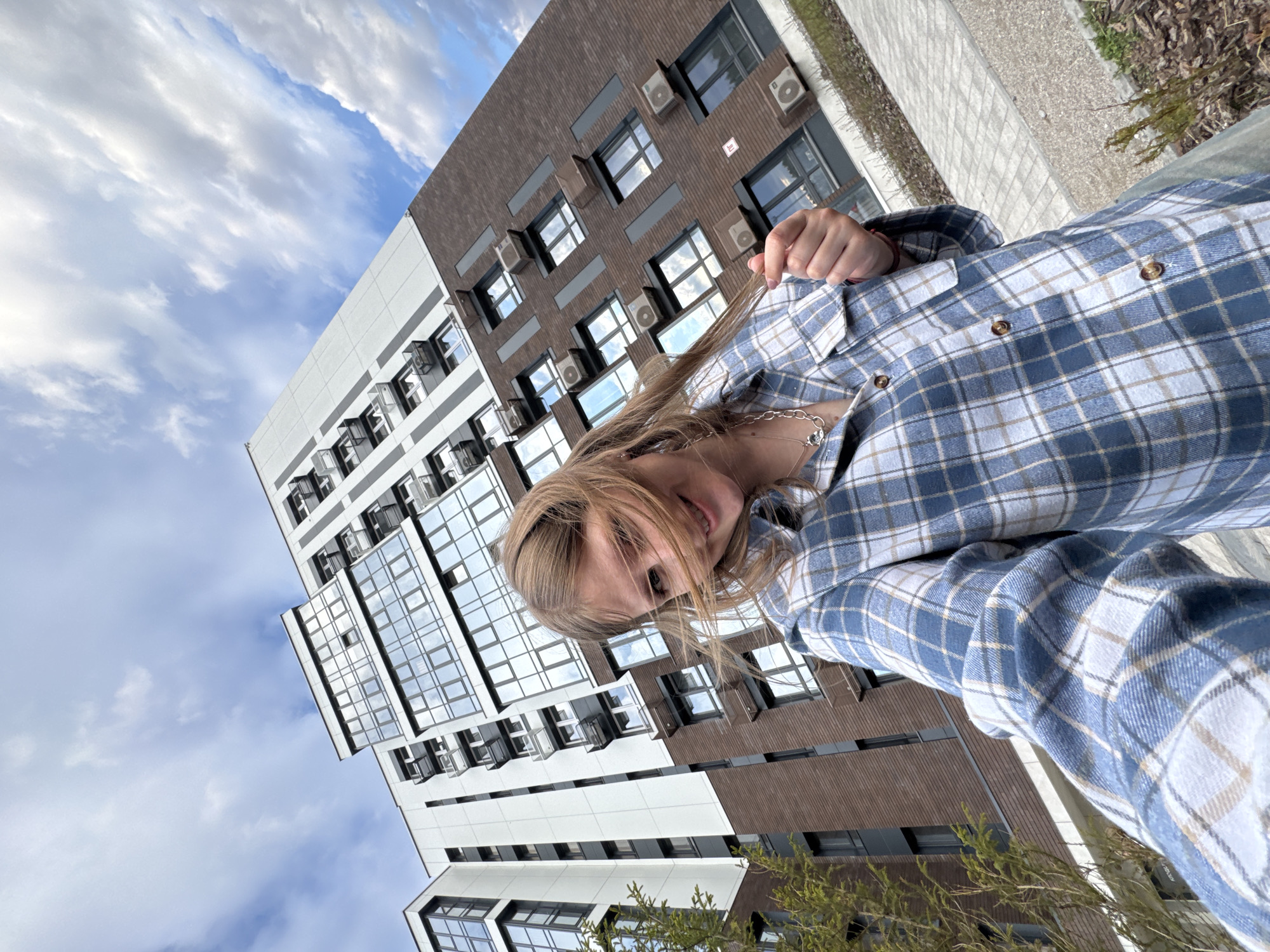 A young woman with long, light blonde hair is featured prominently in a tilted, dynamic shot, likely a selfie. She is smiling softly, with her head slightly tilted to the side, and her hair partially obscures her face. She is wearing a blue, white, and tan plaid button-up shirt and a delicate silver chain necklace with a small charm. Her hand is raised, possibly adjusting her hair or casually posing.
The background consists of modern urban architecture, likely in Perm, Russia. Several multi-story buildings are visible, characterized by a mix of facades: one features a brown, brick-like material, while another is white with dark horizontal stripes. Both buildings have numerous large glass windows reflecting the sky, and many external air conditioning units are mounted below the windows. The sky above is partly cloudy with patches of blue visible, indicating daytime. Some sparse greenery and paved ground are also visible along the bottom and right edge of the frame. The overall mood is casual and bright. A small, red sign with illegible white text is visible on the brown building.