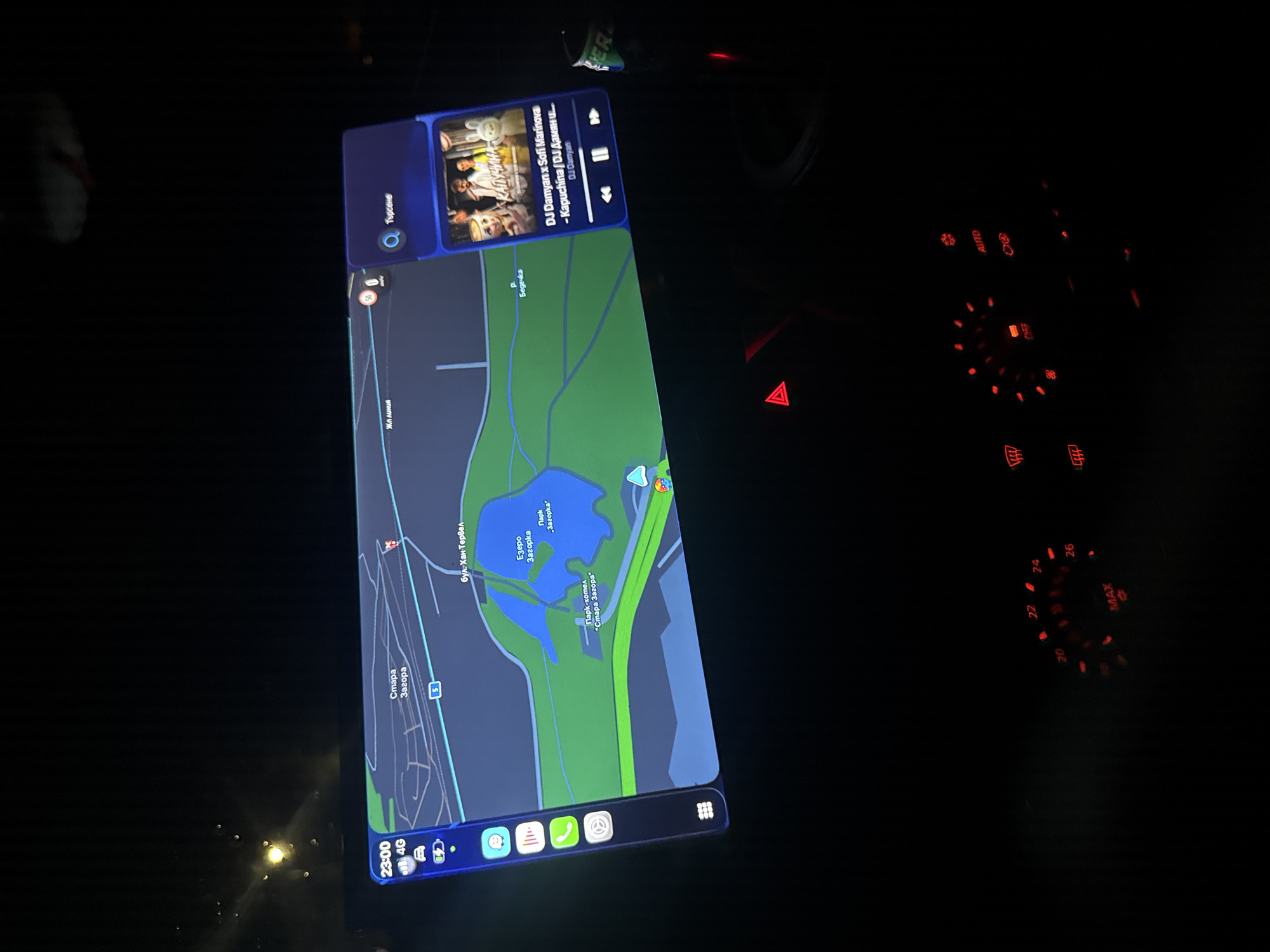 The image shows a dashboard navigation screen displaying a map in the city of Stara Zagora, Bulgaria. The map centers on the area around "Lake Zagorka" and "Zagorka Park." Street "bul. Han Tervel" and "Stara Zagora" are also visible on the screen. At the top of the screen, music is playing by "DJ Damyan x Sofi Marinova." The time displayed is 23:00, and a 4G network connection is active. The hazard lights are flashing. The car's climate control dials are lit up.