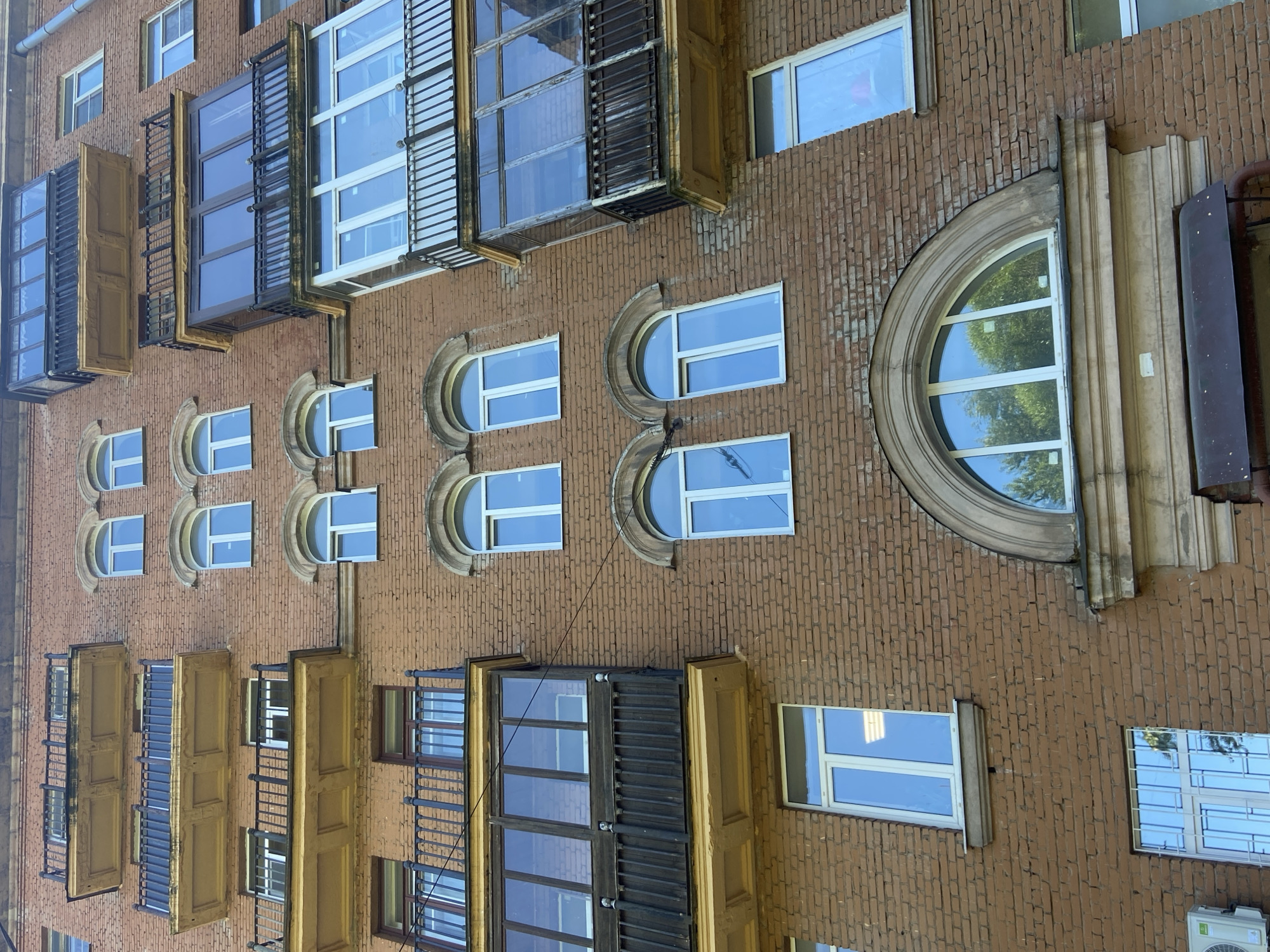 This is a vertical, medium-shot photograph of a brick building's facade in Saint Petersburg, Russia. The building features multiple windows of various shapes and sizes, some with balconies. The windows are mostly rectangular with white frames and appear to be modern replacements in older openings. There are also several rounded arched windows, some with decorative stone surrounds.
The balconies are a mix of open metal railings and enclosed units with wooden shutters. Some shutters are open, revealing the windows behind them, while others are closed. One large, prominent balcony on the right side of the image has a dark, metallic exterior and appears to be enclosed.
The brickwork is a reddish-brown color and shows signs of age and wear. The lighting suggests it is daytime, with sunlight reflecting off the windows. The overall scene depicts an older apartment building undergoing some renovations or having a mix of old and new architectural elements. There is no visible text in the image.