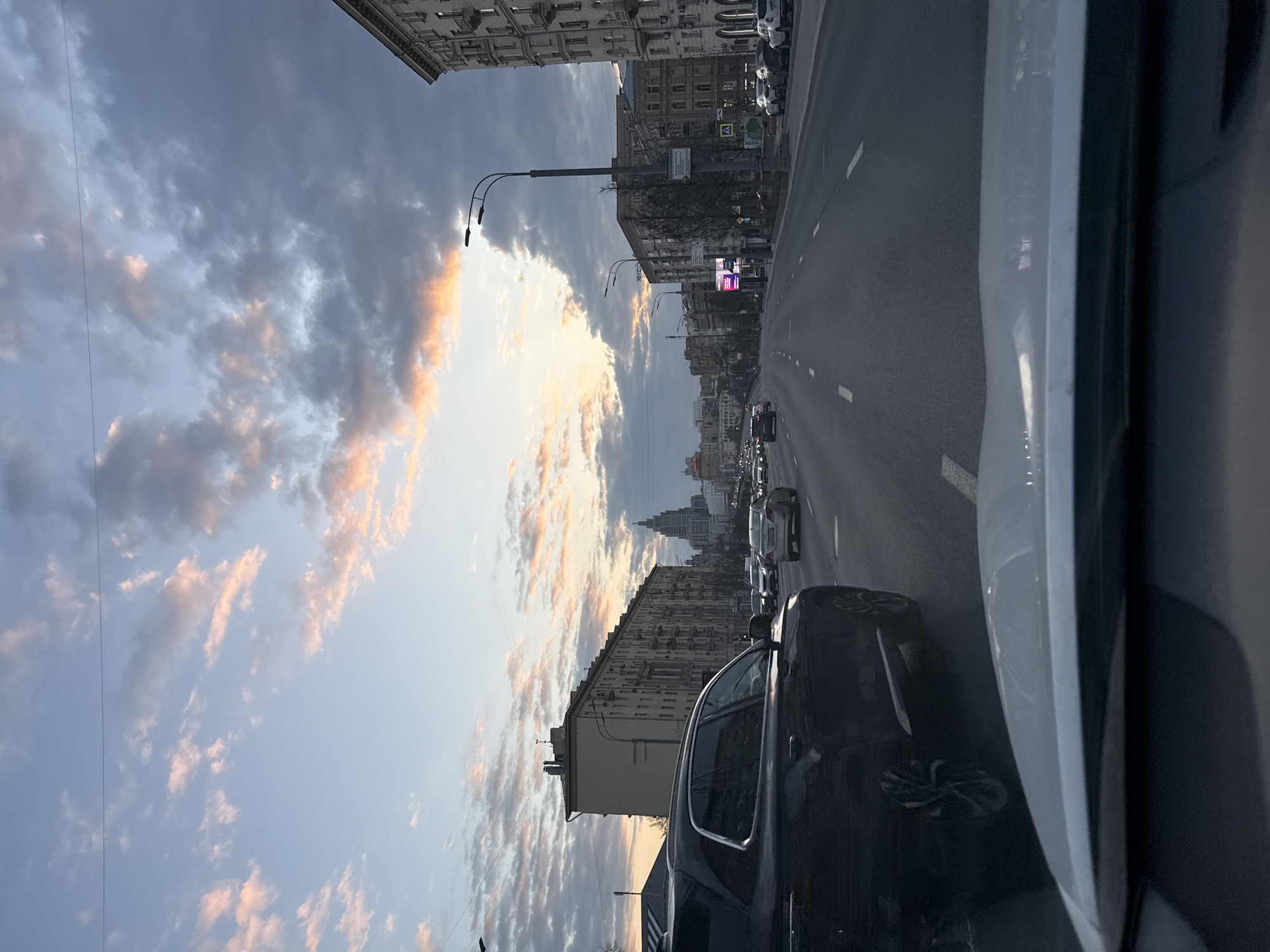 The image shows a view from inside a car, looking out over a city street with multiple lanes of traffic. On either side of the road are tall buildings, suggesting an urban environment. The sky is filled with clouds tinged with pink, indicating it might be sunrise or sunset. Other cars are visible in the street. The location is Moscow, Russia.