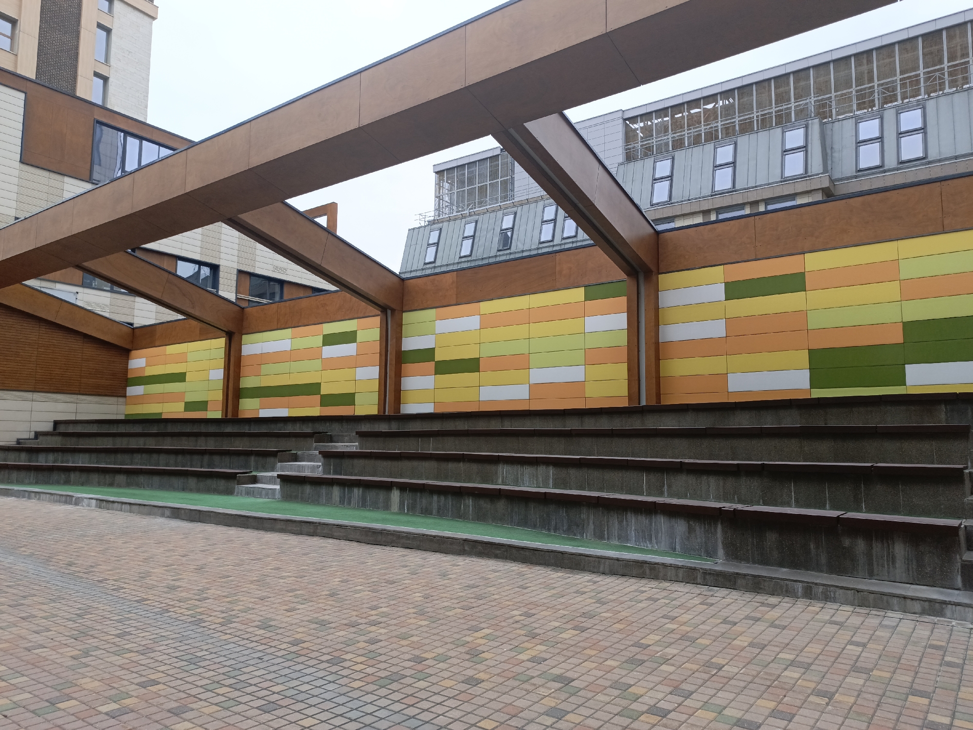 This image depicts a stepped seating area or small amphitheater in what appears to be an urban environment. The location is Moscow, Russia. The seating steps are constructed from concrete and are adorned with strips of green material, possibly artificial turf or some kind of covering, running along the seating surface. The seating backs are segmented into sections, each displaying a pattern of horizontal rectangles in alternating colors such as yellow, green, orange, and white. A large, wooden framework or pergola structure covers the area, creating a sense of enclosure. The flooring in front of the seating is paved with small, multicolored tiles arranged in a grid pattern. The background features buildings of varying heights, including some with modern architectural elements. The weather appears overcast, with a grey sky visible above the structures. No people are present in the scene.