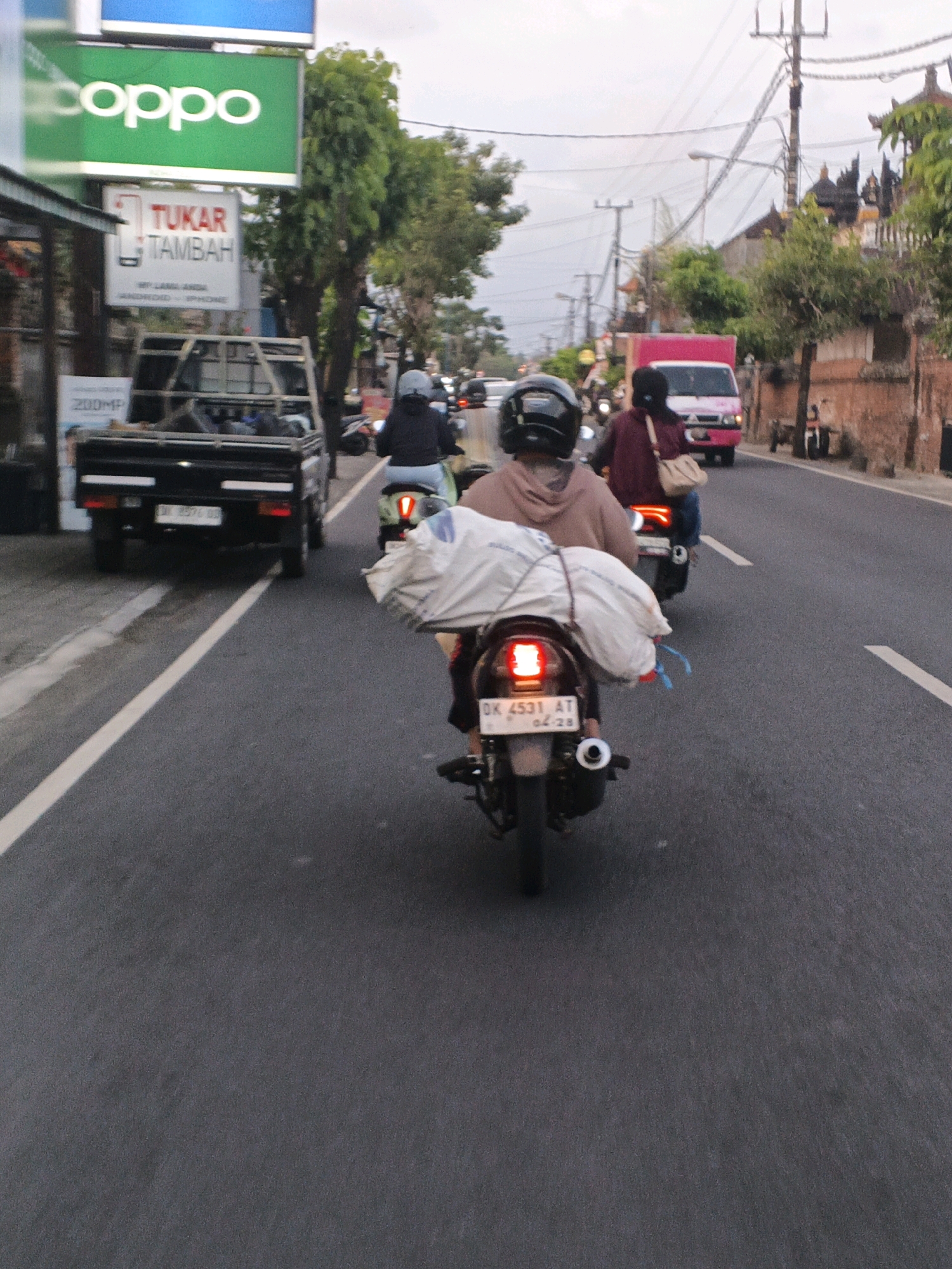 The image depicts a street scene in Denpasar, Indonesia, with multiple motorcycles and a truck on a paved road. In the foreground, two motorcycles are visible. The closest motorcycle has a person wearing a brown hoodie and a black helmet riding it, with a large white sack strapped to the back. The motorcycle's license plate reads "DK 4531 AT 04-28". To the left of this motorcycle, a black truck is parked on the side of the road. Further down the road, another motorcycle is seen from behind, with a person wearing a maroon jacket and a light-colored bag on their shoulder. In the background, a pink truck is driving, and buildings with traditional Balinese architecture are visible along the roadside. Overhead, power lines crisscross the sky. A green sign with the word "coppo" in white letters is also visible on a building to the left. The overall atmosphere suggests a typical day of traffic and commerce in the city.
