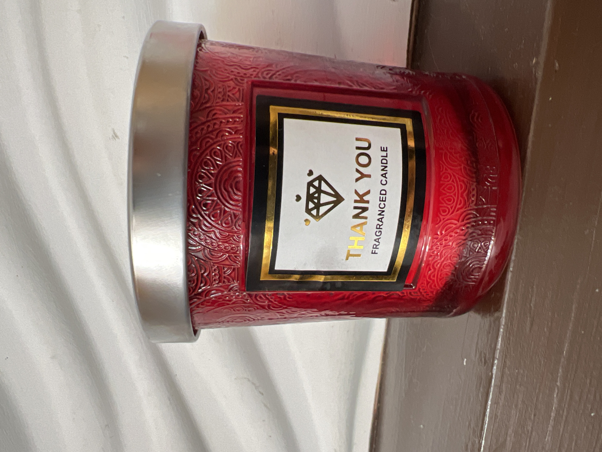 A red, textured glass candle jar with a silver lid sits on a wooden surface. The jar has an ornate, embossed pattern around its circumference. A label with a black border and gold trim is affixed to the front of the jar. The label displays the text "THANK YOU" in large, gold, sans-serif font. Below "THANK YOU" is an image of a gold diamond. Underneath the diamond, the text "FRAGRANCED CANDLE" is printed in smaller, gold, sans-serif font. The candle appears to be illuminated from above, casting a soft light on the surrounding surfaces. The background is a textured white surface on the left and a dark brown wooden surface on the right. The overall impression is one of a decorative and aromatic item.