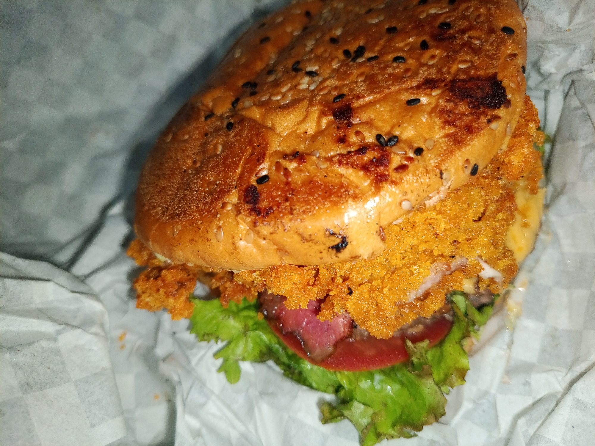 The image shows a hamburger in Tinaquillo, Venezuela. The burger has a sesame seed bun, lettuce, tomato, and a breaded patty with orange breading. The burger is placed on white checkered wax paper. The lighting is dim, and the focus is on the burger.