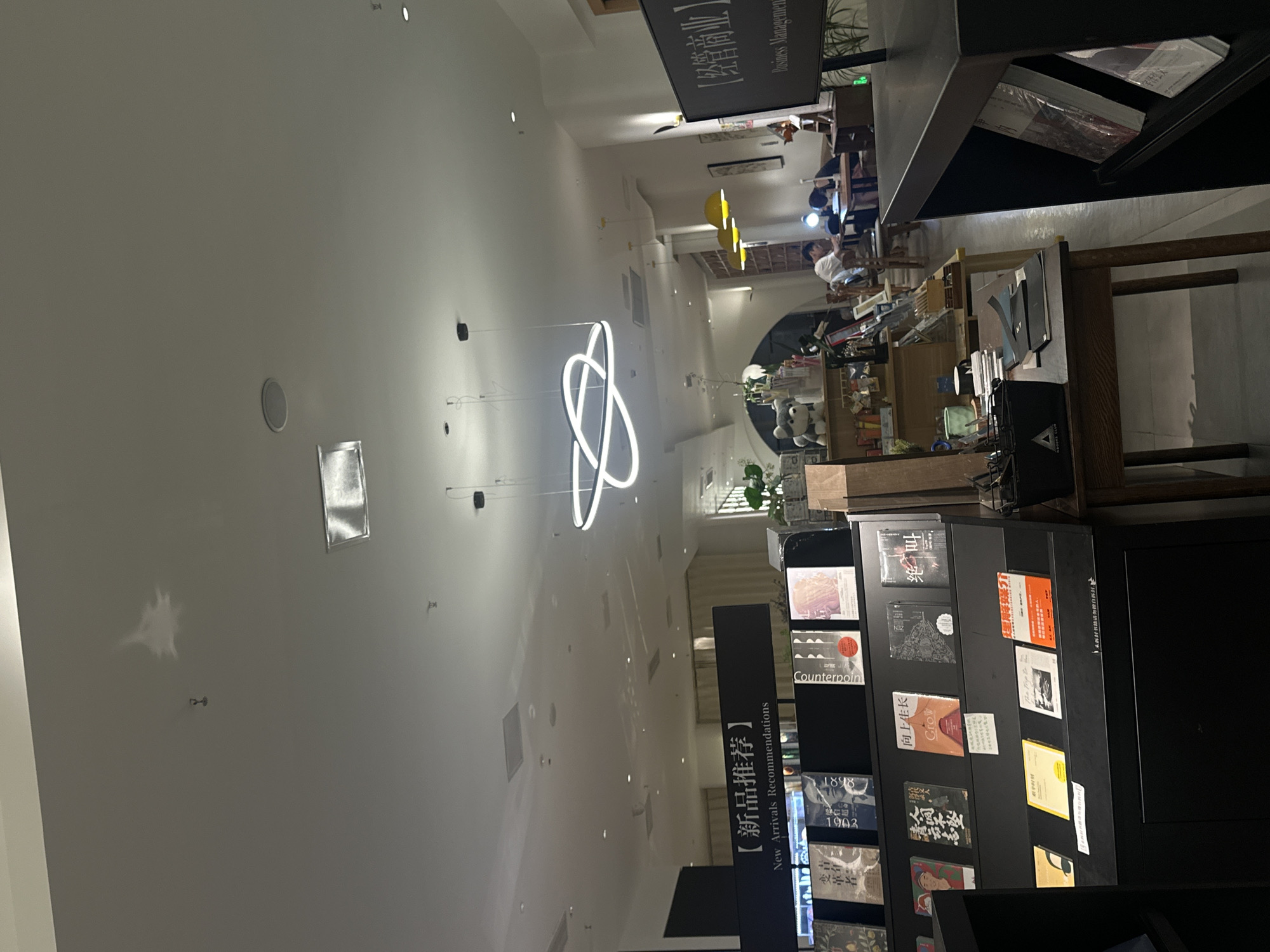The image depicts the interior of a modern bookstore or cultural space, likely located in Chongqing High-tech Zone, China. The scene has a high, white ceiling with recessed lighting and circular speakers, creating a spacious and brightly lit atmosphere.
In the foreground, on the right side of the frame, a black, multi-tiered display shelf is prominently featured, showcasing various books. Text on this display reads "【新品推荐】New Arrivals Recommendations". Visible book titles include "Counterpoint," "1898," "1903," "向上生长 Grow," "绝叫," "人间不坠," and "变革者."
Further into the midground and background, an area with tables and chairs is visible, where several people are seated. They appear to be reading, working, or quietly conversing, suggesting a calm and studious environment. This seating area is illuminated by three yellow dome-shaped pendant lights, contributing to a warm ambiance. A large black sign hanging in the upper-right background carries the text "【经管商业】Business Management," indicating a specific section or theme within the venue.
A striking white neon light fixture, designed as an abstract, intertwined loop, hangs centrally from the high ceiling, serving as a modern decorative element. Other objects visible include additional shelving in the background, a stuffed teddy bear on a shelf, and various small decor items. The overall design emphasizes contemporary aesthetics with a focus on books and a comfortable space for visitors.