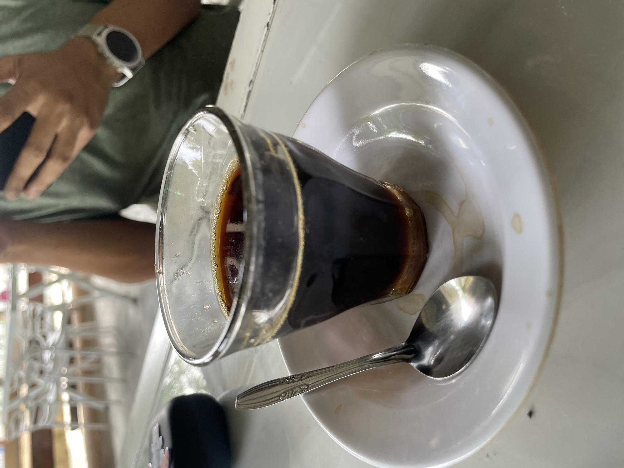 A close-up shot focuses on a glass of dark coffee, placed on a white saucer with a spoon resting to its left. The coffee has a frothy, light brown layer on top and appears to be a concentrated brew, possibly Indonesian coffee. The glass itself is clear and has a slightly flared rim. The saucer has a subtle lip and shows a few faint brown stains, suggesting it has been used. The spoon is made of metal and has a decorative pattern on its handle. In the background, a person is visible, wearing a green t-shirt and a smartwatch on their left wrist, and holding a dark-colored smartphone. The setting appears to be an outdoor cafe or a similar establishment, with blurred white tables and chairs in the distance, suggesting a public space in Pasar Paloh, Indonesia.