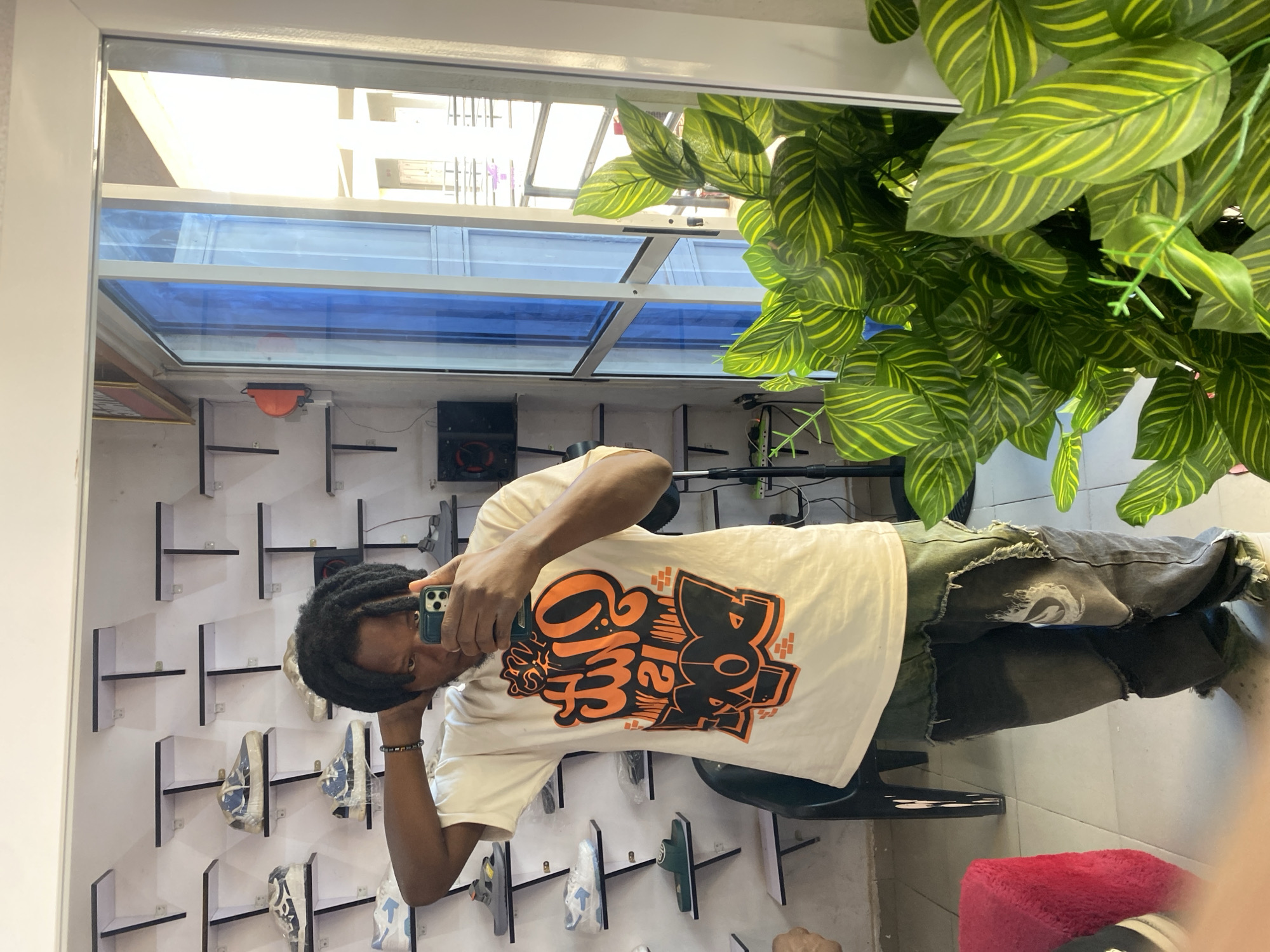 A young man with dreadlocks takes a mirror selfie in a retail space, likely a shoe store, located in Jos, Nigeria. He is wearing a cream-colored t-shirt with an orange and black graphic design and distressed dark wash jeans. He holds a smartphone in his right hand, framing the shot. The background features shelves displaying various sneakers and footwear. A tall, potted plant with vibrant green and yellow striped leaves is positioned to the right of the frame. In the background, a blue-tinted glass door is visible, revealing glimpses of the exterior.
