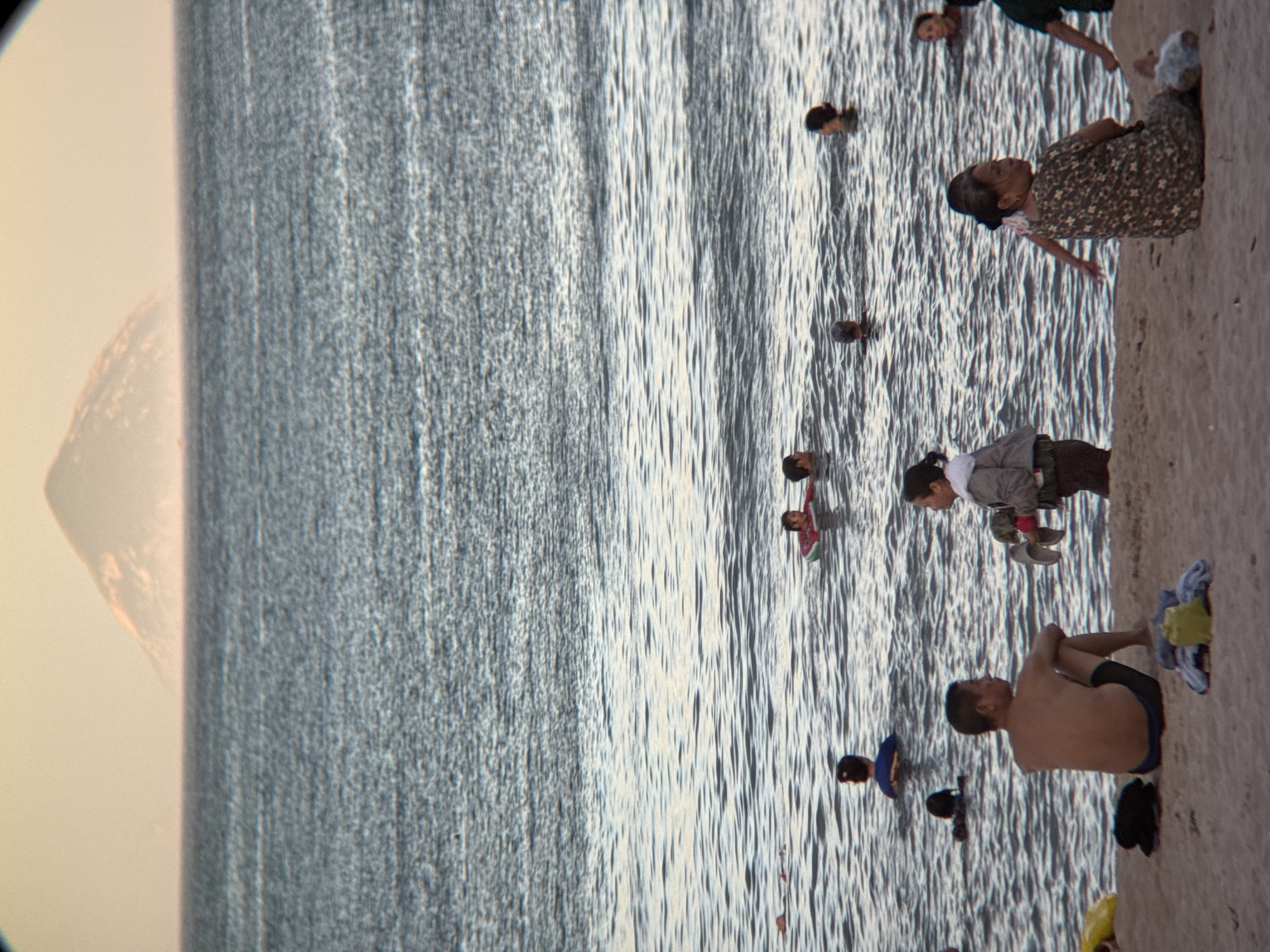The image depicts a beach scene in Nha Trang, Vietnam, with the ocean stretching towards a hazy, triangular-shaped island in the distance. The sky is a pale, warm color, suggesting it might be early morning or late afternoon.
In the foreground, the sandy beach is sparsely populated. A man in blue swim trunks is seated on the sand, facing away from the camera. To his right, a woman in a patterned dress sits on the sand, looking to her left. Next to her, a young girl in a light-colored jacket is standing, holding what appears to be a pair of shoes or sandals. Various items of clothing and a yellow bag are scattered on the sand near the seated individuals.
In the water, several people are visible. Some are swimming, while others are wading. A person in a red inner tube is in the middle ground. The water is calm with gentle ripples. The overall atmosphere appears relaxed, with people enjoying a day at the beach.