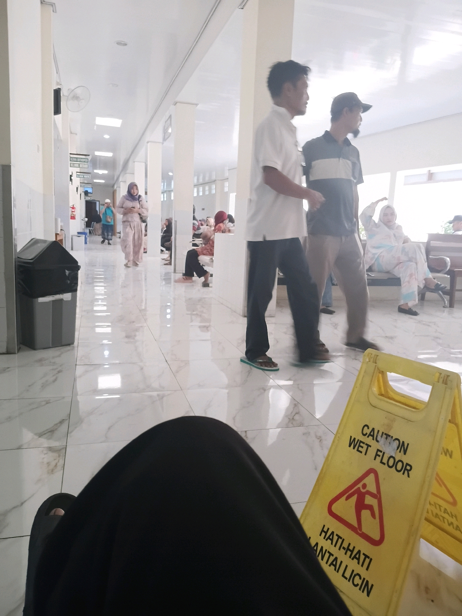 The image depicts a long, brightly lit hallway, likely within a public building such as a hospital or government office, in Purwodadi, Indonesia. The floor is highly polished white tile with a subtle marble pattern, reflecting the overhead fluorescent lighting. Several people are present, either standing or sitting, engaged in various activities. In the foreground, a black garment, possibly a skirt or robe, obscures the lower portion of the frame, with a foot wearing a black sandal visible. To the right, a yellow "Caution: Wet Floor" sign with a pictogram of a person slipping is prominently displayed, suggesting a recent cleaning or a spill. The sign also has text in Indonesian: "HATI-HATI LANTAI LICIN" (Be careful, slippery floor).
Further down the hallway, a woman in a light purple hijab and matching outfit walks away from the camera, holding a phone in her right hand. Two men are also visible: one in a white short-sleeved shirt and dark pants, and another in a dark polo shirt with a horizontal stripe and a baseball cap. They appear to be in conversation or observing something. In the background, other individuals are seated on a bench, and a large grey trash bin with a black lid is positioned against a white wall to the left. The overall impression is one of a busy, functional indoor public space.