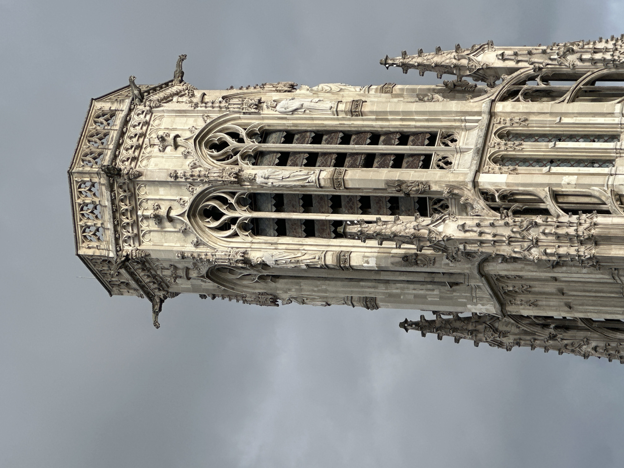 The image shows a close-up of a section of the Saint-Jacques Tower (Tour Saint-Jacques) in Paris, France. The tower is depicted in a vertical orientation against a cloudy sky. The architectural details are highly visible, including intricate carvings, pointed arches, and multiple layers of ornamentation. Gargoyles and other decorative figures are present. The tower appears to be constructed from light-colored stone, likely limestone. No people are visible in the image.