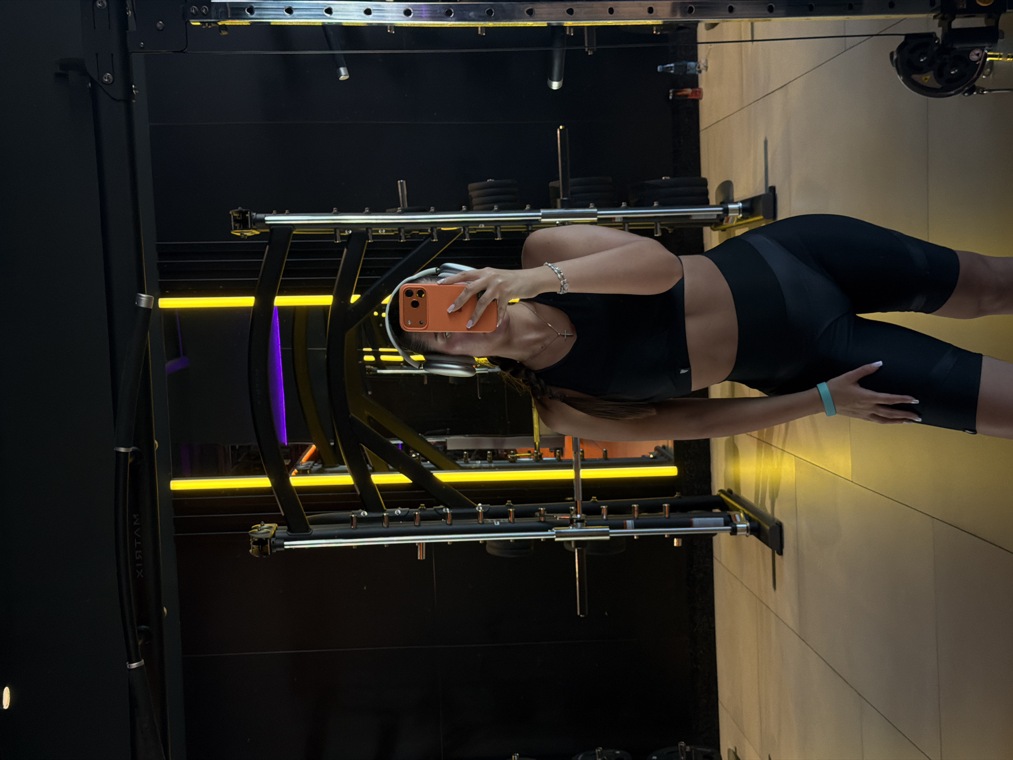 A young woman stands in a gym in Krasnoyarsk, Russia, posing for a selfie in a mirror. She is wearing a black sports bra and black cycling shorts. She is holding an orange phone in her left hand and has white headphones on. Her hair is in a braid. The gym equipment is visible in the background, including a Smith machine with yellow and purple lights and a cable machine. The floor is tiled.