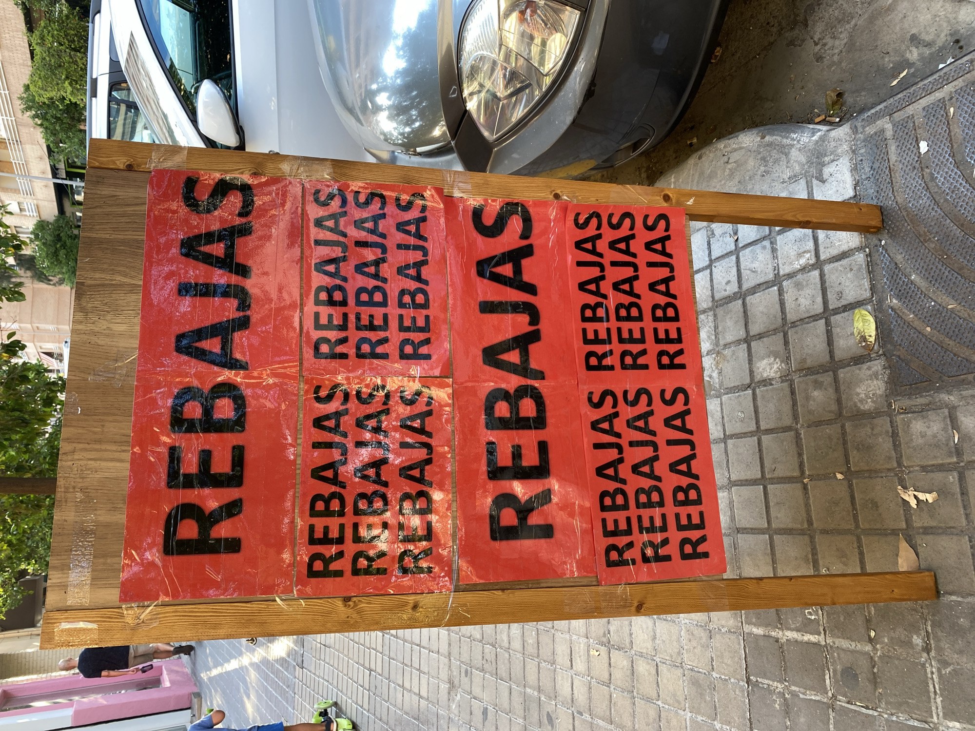 A group of red signs with black text reading "REBAJAS" (Spanish for "sales") are displayed on a wooden stand. The signs are rectangular and appear to be made of a shiny material, possibly plastic or laminated paper. The text is large and bold, with "REBAJAS" repeated multiple times on each sign. Some of the signs are partially covered by clear plastic sheeting, which creates a wrinkled effect.
The wooden stand is made of two planks, one horizontal and one vertical, supporting the signs. The stand is positioned on a paved area with square tiles, and a textured rubber mat is visible on the right side of the frame. In the background, a white car is parked, with its headlight and part of the grille visible. Trees and a building can be seen in the upper left corner of the image, suggesting an outdoor urban setting.
The scene appears to be during daylight, with shadows cast on the ground. The overall impression is that of a commercial display, likely advertising a sale event. No people are clearly visible in the frame, though a glimpse of a person's legs in shorts and sandals can be seen in the lower portion of the image, suggesting the photo was taken by someone standing or walking nearby. The lighting and general atmosphere suggest a pleasant day, possibly in a Spanish city like València, given the "REBAJAS" text and the architectural cues.