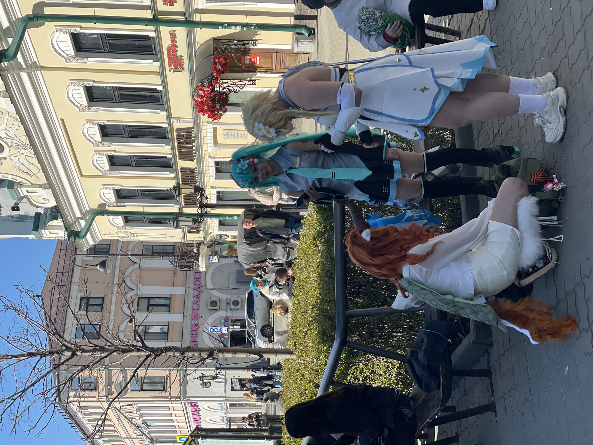 The image captures a street scene in Kazan, Russia. Three individuals are in cosplay, sitting on or next to a low metal fence that borders a raised planting bed filled with green shrubs. One individual has orange hair, a white outfit, and a bushy orange tail. Another cosplayer is wearing a blue-green wig, a grey top with a blue tie, and a black skirt. A third cosplayer has blond hair with white flowers and a white dress with blue trim. In the background, there is a crowd of people walking and standing on the sidewalk. Behind the crowd, buildings line the street, with a mix of architectural styles. One building has a sign with the Russian words "ЗАМОК СЛАДОС" in pink neon letters. The sky is blue and mostly clear. A bare tree stands to the left, without leaves. The pavement is patterned with grey bricks. Red flowers adorn a decorative balcony on one of the buildings.