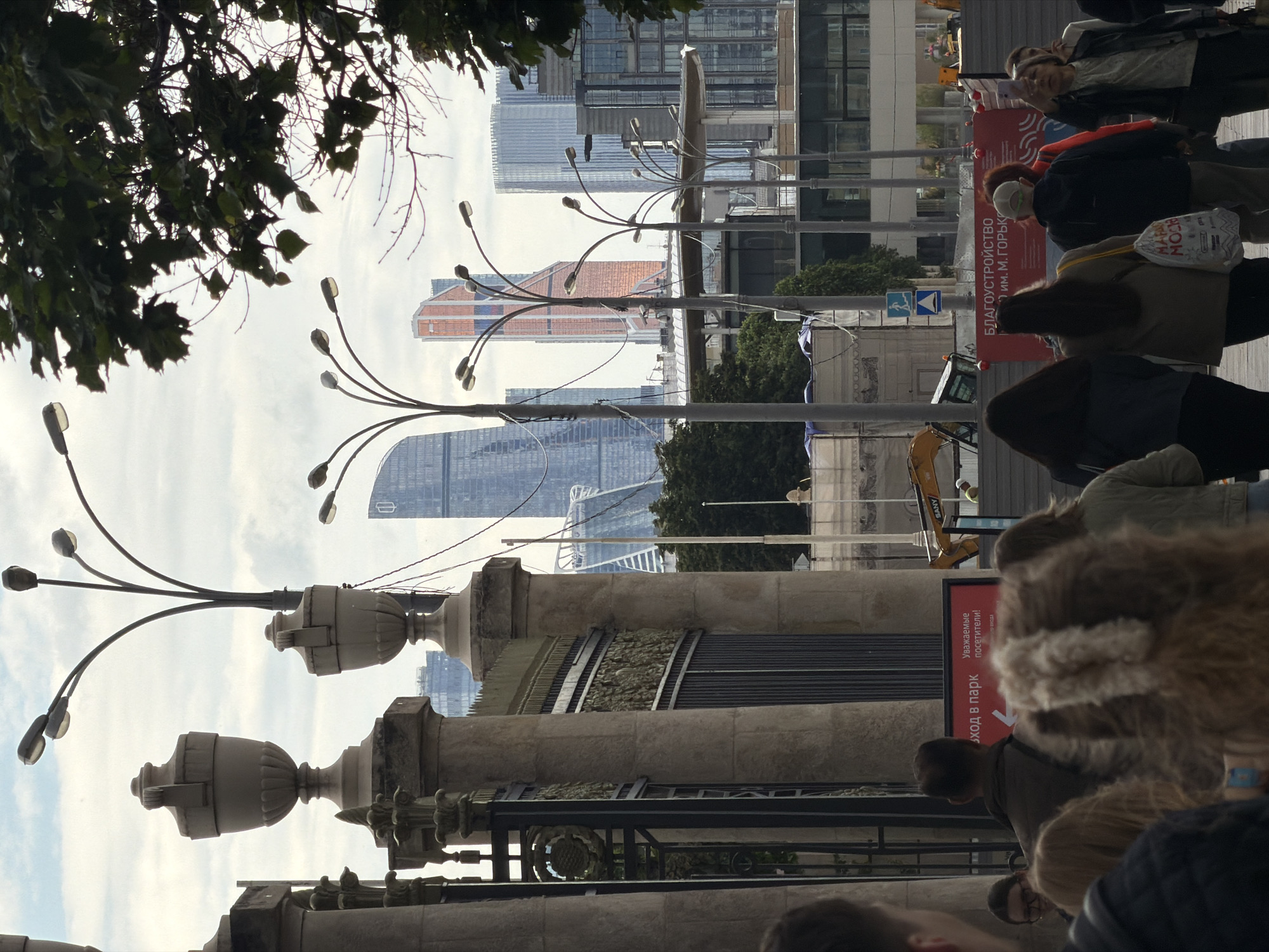 Here is a detailed description of the image:
The image appears to be taken from a low angle, looking up and across a street scene in Moscow, Russia. The foreground shows a stone or concrete structure, possibly a bridge or a park boundary. It features ornate urn-like decorations and metal railings.
Beyond the structure is a pedestrian walkway with people walking by. A red sign with white Cyrillic text is visible, likely advertising an event or directing people to a location. A yellow excavator is also visible, suggesting construction work nearby.
In the background, modern high-rise buildings dominate the skyline, characteristic of the Moscow International Business Center (Moscow City) financial district. The sky is overcast, providing a soft, diffused light.
The image is somewhat obstructed by branches and leaves in the upper left, framing the shot. The composition suggests a dynamic urban environment, with a mix of old and new architecture, public spaces, and ongoing development.