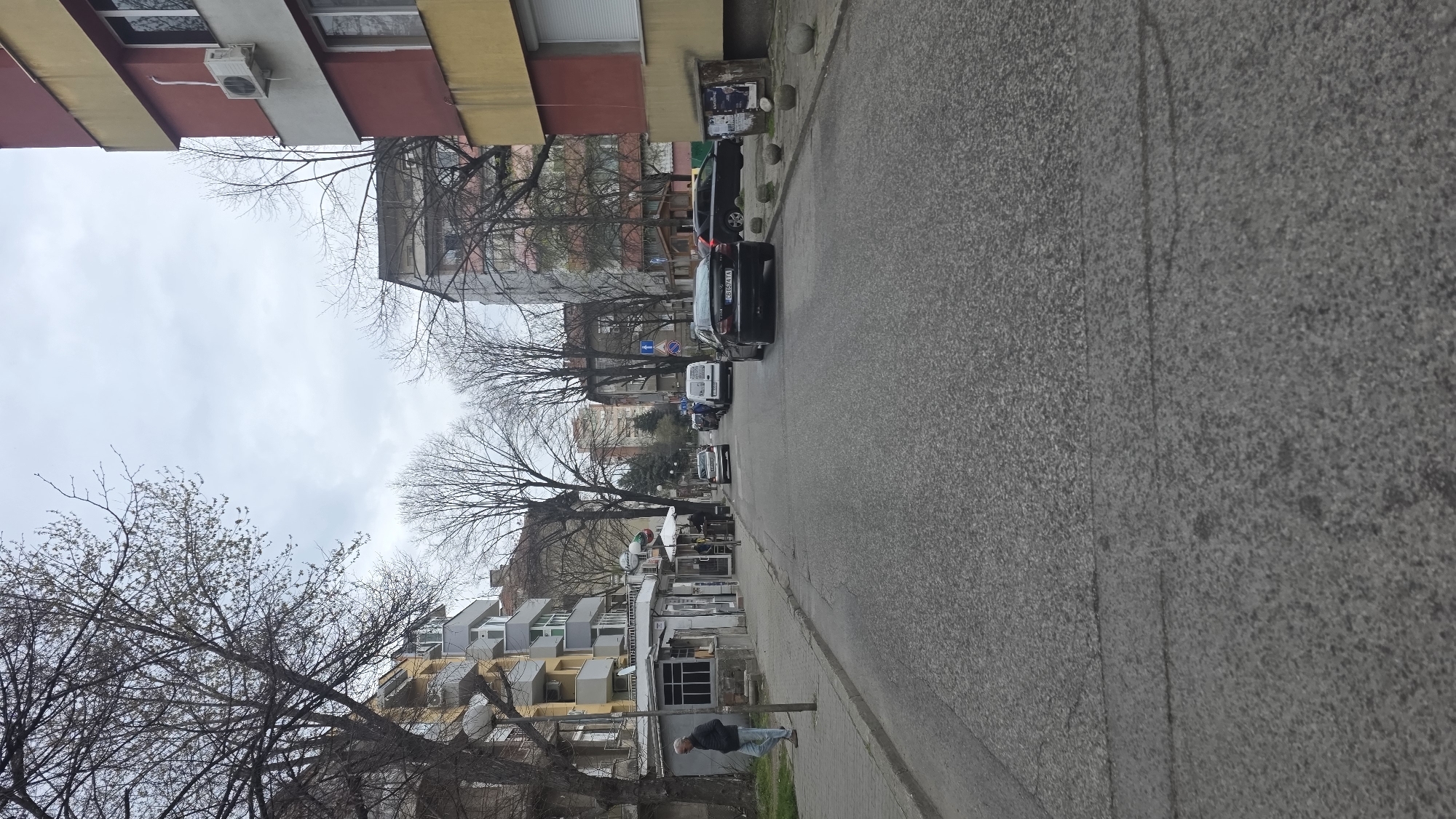 The image shows a street in Blagoevgrad, Bulgaria. Buildings line the street on both sides, a mix of residential apartments with yellow and red accents and commercial properties. Trees with bare branches are visible, creating a bare canopy above. Cars are parked along the right side of the street, and one vehicle is moving in the street. A pedestrian is walking along the sidewalk on the right. The street is paved with asphalt, and the sky is overcast.