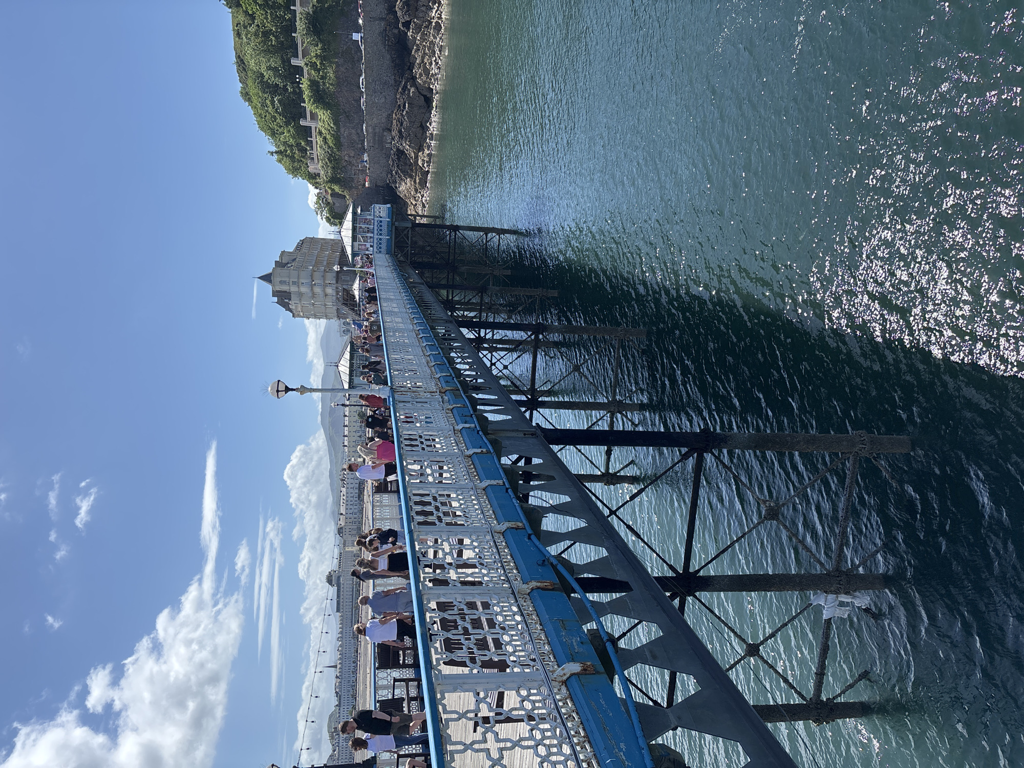 The image shows the Llandudno Pier, located in Llandudno, Wales, United Kingdom. It is a long pier extending out into the sea. People are walking along the pier, looking out at the water. The pier's structure is visible beneath the walkway, and there is a building at the end of the pier. The sea is calm and reflects the sunlight. The sky is blue with scattered clouds. A cliff face is visible on the left side of the image.
