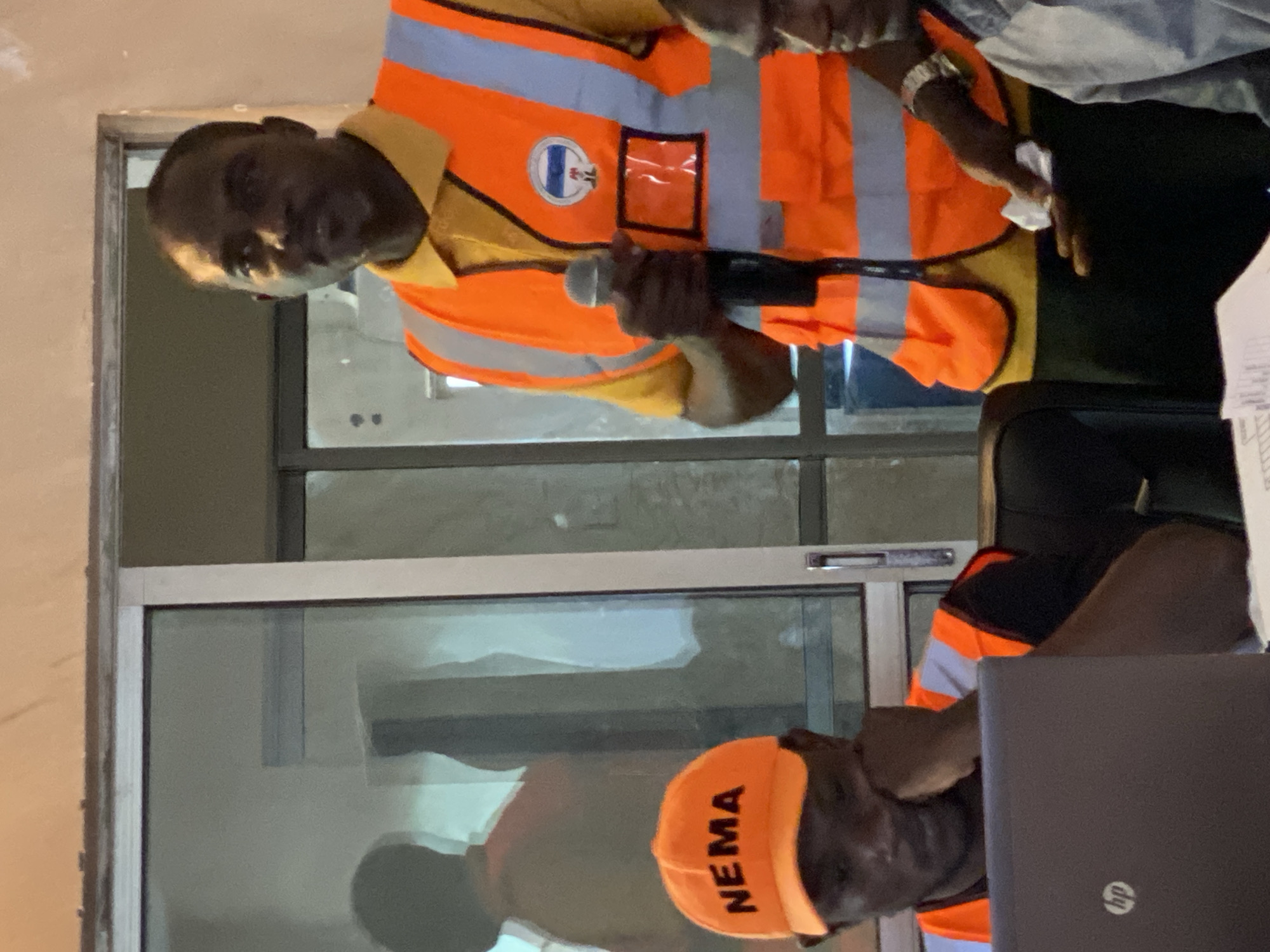 In Unguwar Sarki, Nigeria, a man in a bright orange reflective vest stands near a window. The vest has silver reflective strips and a circular emblem with blue and white elements. He holds a microphone in his right hand and wears a yellow collared shirt underneath the vest. To his left, another man is seated, wearing an orange cap with the word "NEMA" in black lettering and a black t-shirt. A laptop is in front of him, and his left hand rests on his chin. The environment appears to be indoors, with reflective surfaces visible through the window.