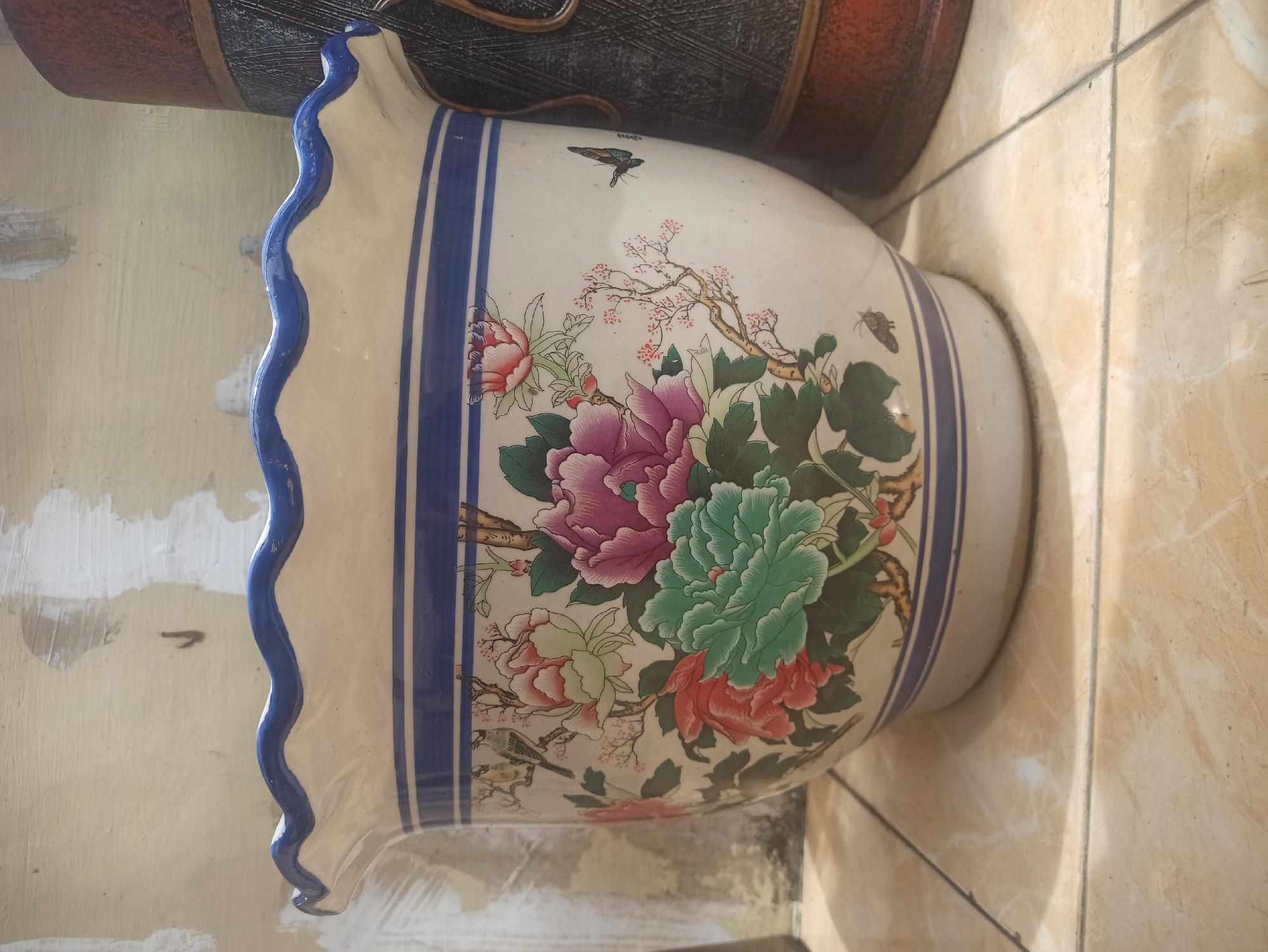 A wide, ornate ceramic planter sits on a tiled floor. The planter is cream-colored with a scalloped blue rim and two horizontal blue bands around its body. It is decorated with a detailed, colorful floral motif featuring large peonies in shades of pink, purple, and green, as well as smaller pink blossoms and green leaves. Two small birds are depicted on a branch on the left side of the planter, and a butterfly is shown on the right. The background appears to be a textured wall with patches of white paint. To the right of the planter, a portion of a dark, textured cylindrical object, possibly another vase or container, is visible. The lighting suggests an indoor setting, possibly a home or shop. The scene is static, with no people or animals actively interacting.