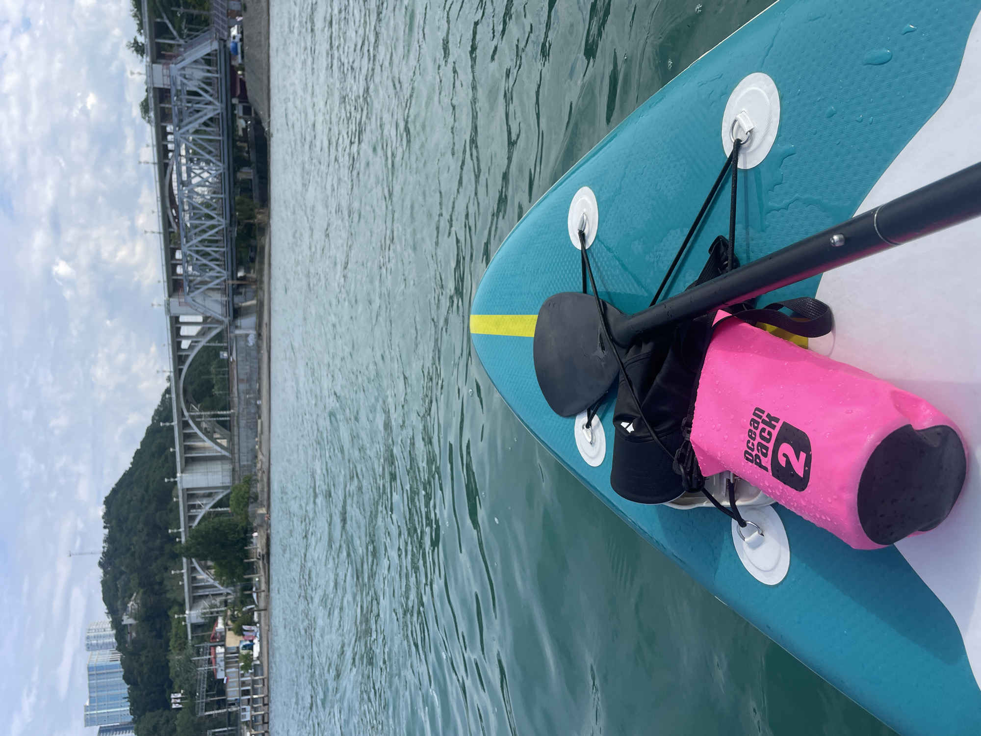The image shows a close-up view of the front of a teal-colored stand-up paddleboard on the water, with a paddle resting on it. A bright pink dry bag labeled "Ocean Pack 2" and a black cap are also on the paddleboard. In the background, a large arched bridge and a truss bridge are visible, spanning across a body of water with buildings and trees on the shoreline. This scene is located in the city of Sochi, Russia, likely near the coastal area where these bridges are prominent landmarks. The water appears calm, with gentle ripples, and the sky is partly cloudy.
