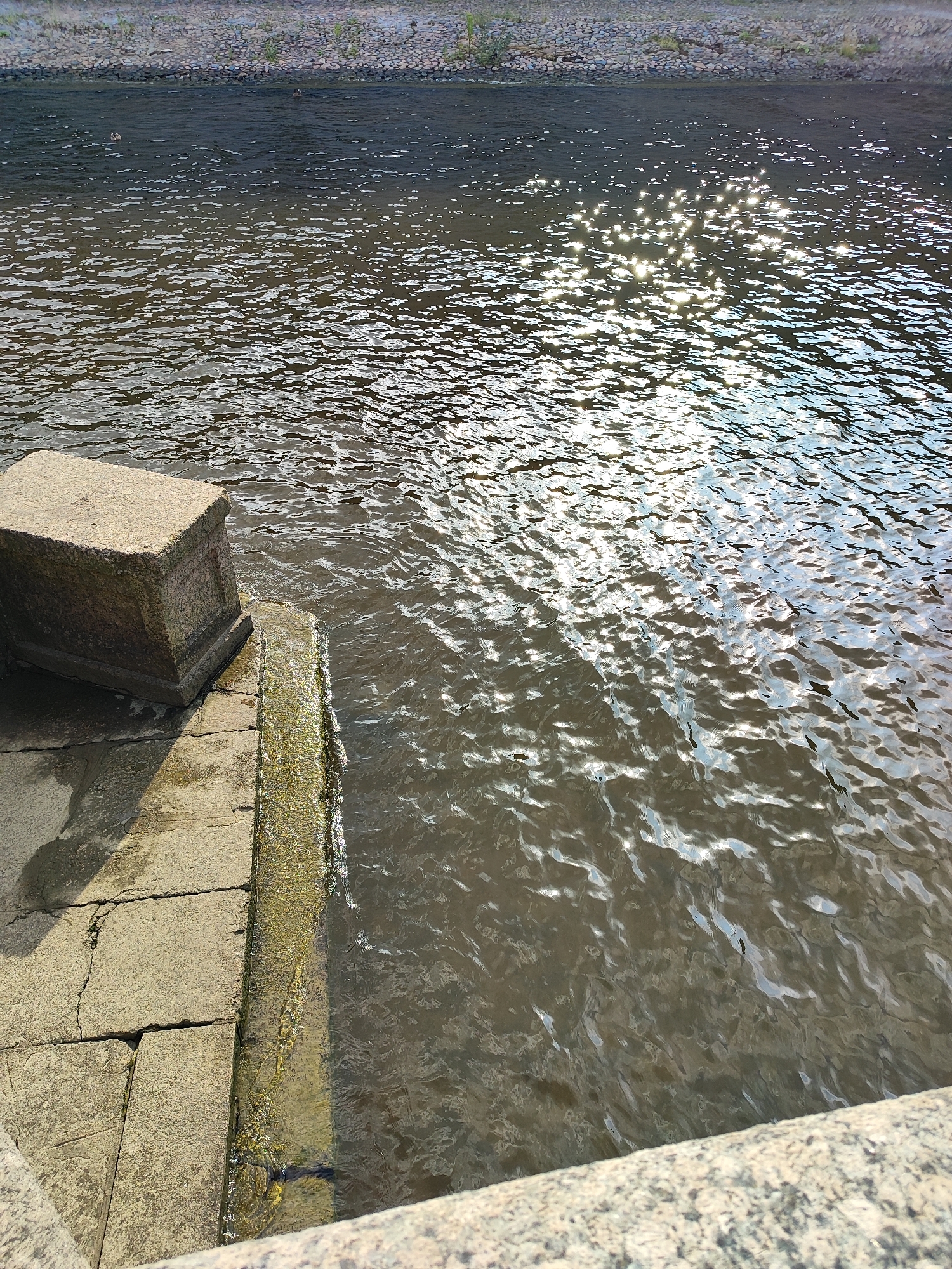 The image shows a canal with dappled sunlight reflecting off the rippling surface. On the left, a stone structure, possibly a bollard or a decorative element, sits on a paved embankment. The embankment is made of large, cracked stone slabs. A narrow strip of darker stone or concrete runs along the edge of the water, covered in some green growth. The far bank of the canal is visible, composed of a layered stone wall topped with dry grass and a few small plants. The water appears slightly murky. The scene suggests a daytime setting in an urban environment, likely a canal or river in Saint Petersburg, Russia, given the architectural style of the embankment. There are no people visible in the image.