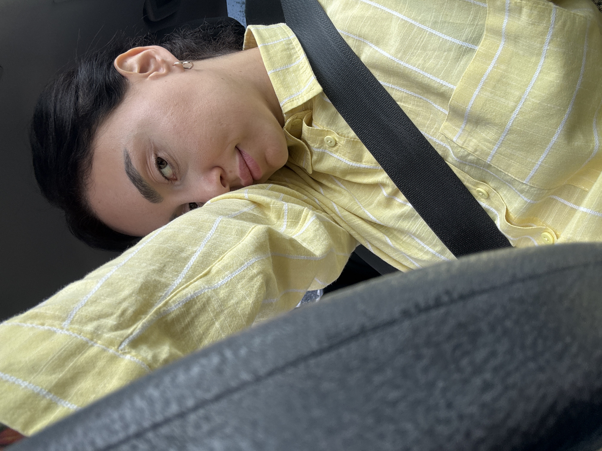 A person with black hair is wearing a yellow and white striped shirt and a black seatbelt, leaning their head on the black leather of what appears to be a car seat. The image is taken from a close angle, possibly inside a car. The location, Tbilisi, Georgia, is not discernible from the image.