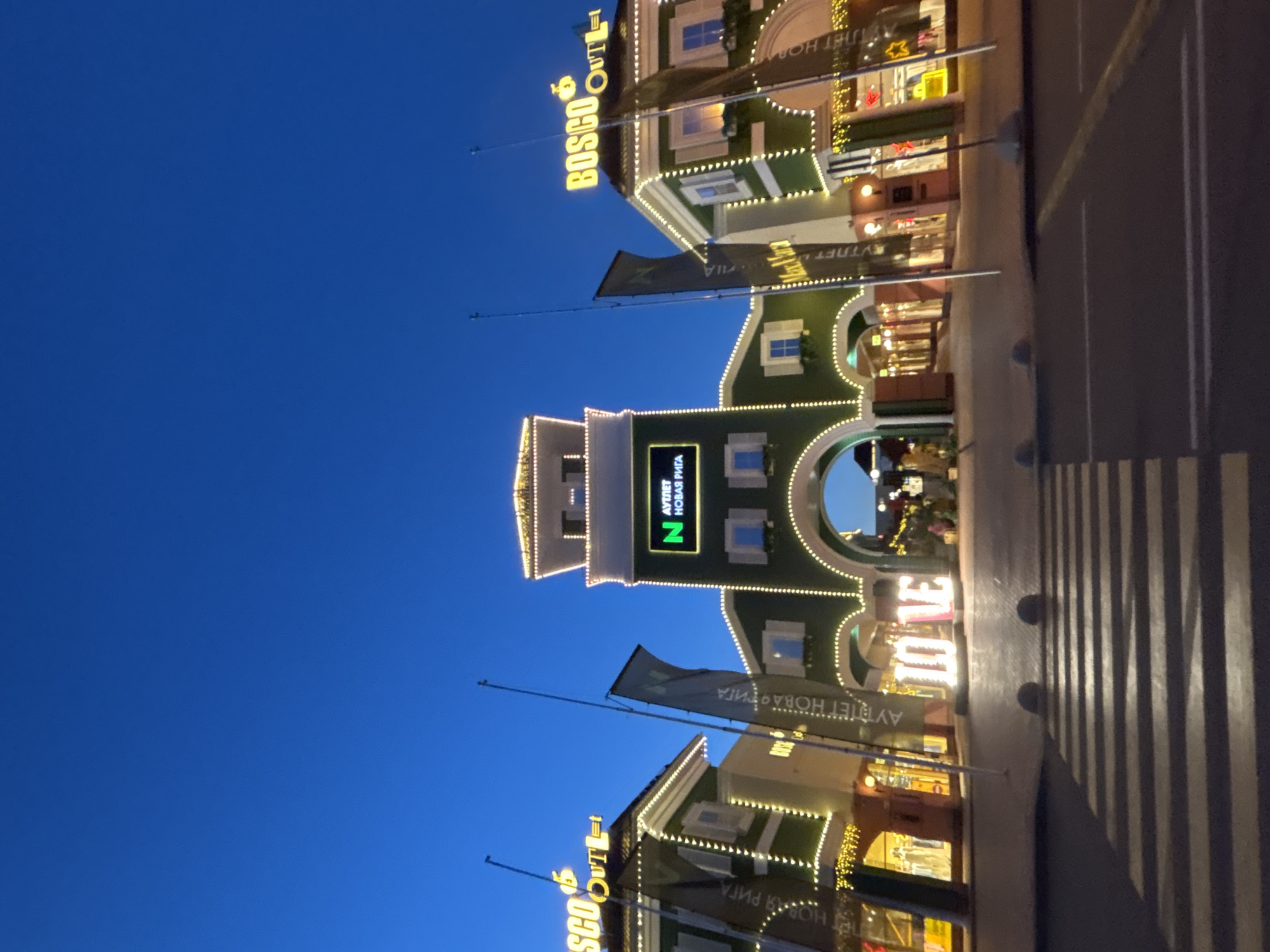 The image shows the Bosco Outlet in Pokrovskoe, Russia, at dusk. The outlet is decorated with numerous lights, highlighting the architecture of the buildings and the "LOVE" sign in the foreground. The sky is a deep blue, indicating the transition from day to night. In the foreground, a pedestrian crossing is visible. There are no people clearly visible in the image.