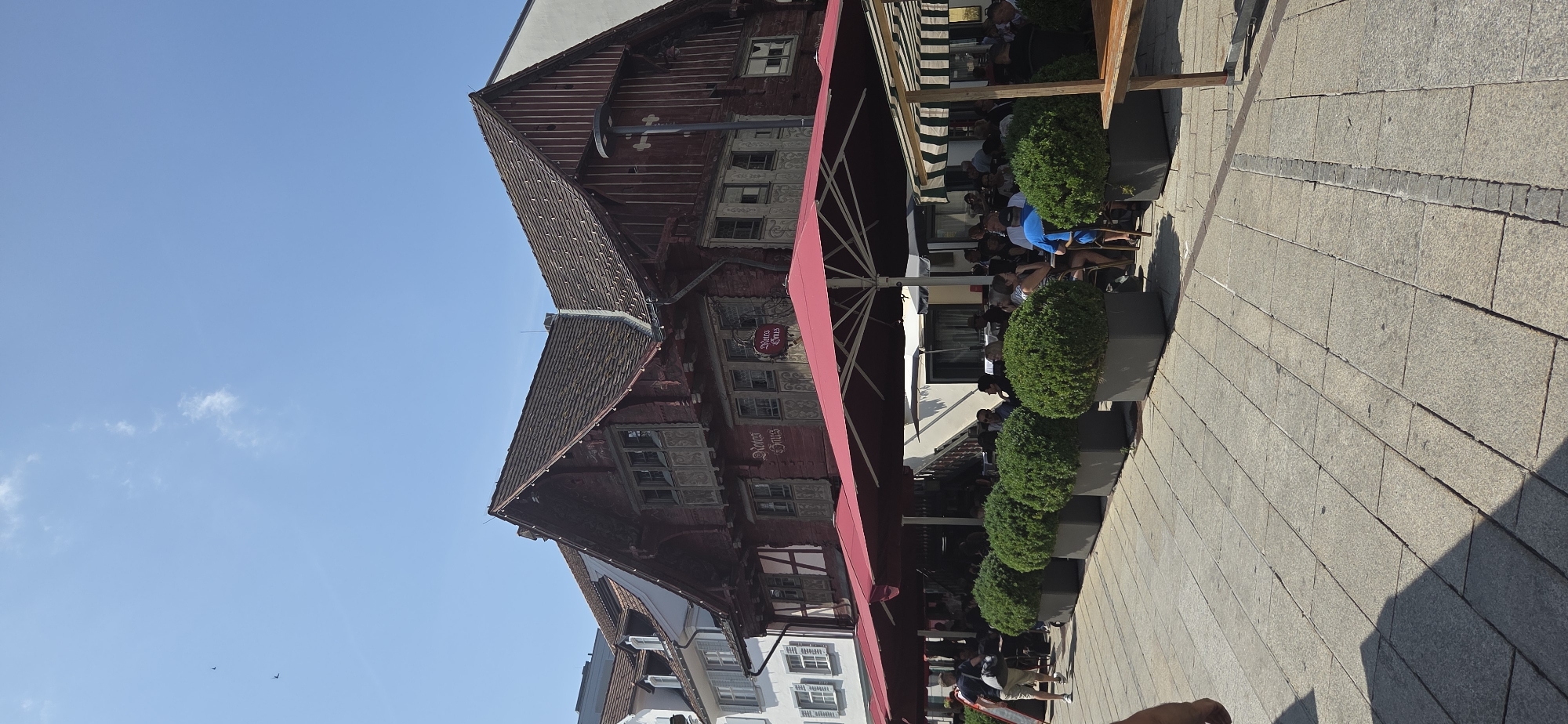 The image shows an outdoor seating area of a restaurant in the city of Dornbirn, Austria. The restaurant, identified by signs as "Rotes Haus," is housed in a traditional red wooden building with a steeply pitched roof. Red umbrellas are set up to provide shade for tables where patrons are seated. In front of the restaurant, several large rectangular planters contain manicured green bushes. The ground is paved with large square stones. Several people are visible sitting at tables and walking around the area. The sky is clear and blue with a few scattered clouds, and the sun is shining brightly, casting shadows on the pavement. The overall scene depicts a pleasant, sunny day in a European city, likely during the daytime given the lighting.