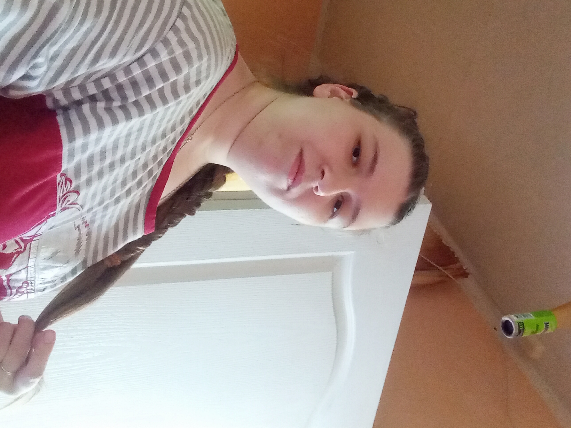 A medium shot captures a young woman indoors, facing forward and slightly to her left, with her right hand holding a strand of her braided hair. She wears a grey and white striped t-shirt with a red panel at the bottom featuring a white floral design. A thin necklace is visible around her neck. The background consists of a pale orange wall on the left, a white door on the right, and a beige ceiling above, where a bright green object, possibly a roll of tape or a small can, is affixed. The lighting is somewhat dim, with a strong light source to the right, creating a bright highlight on the door. The image is taken from a low angle, looking up towards the ceiling. This appears to be a residential interior, potentially in Kitskany, Moldova.