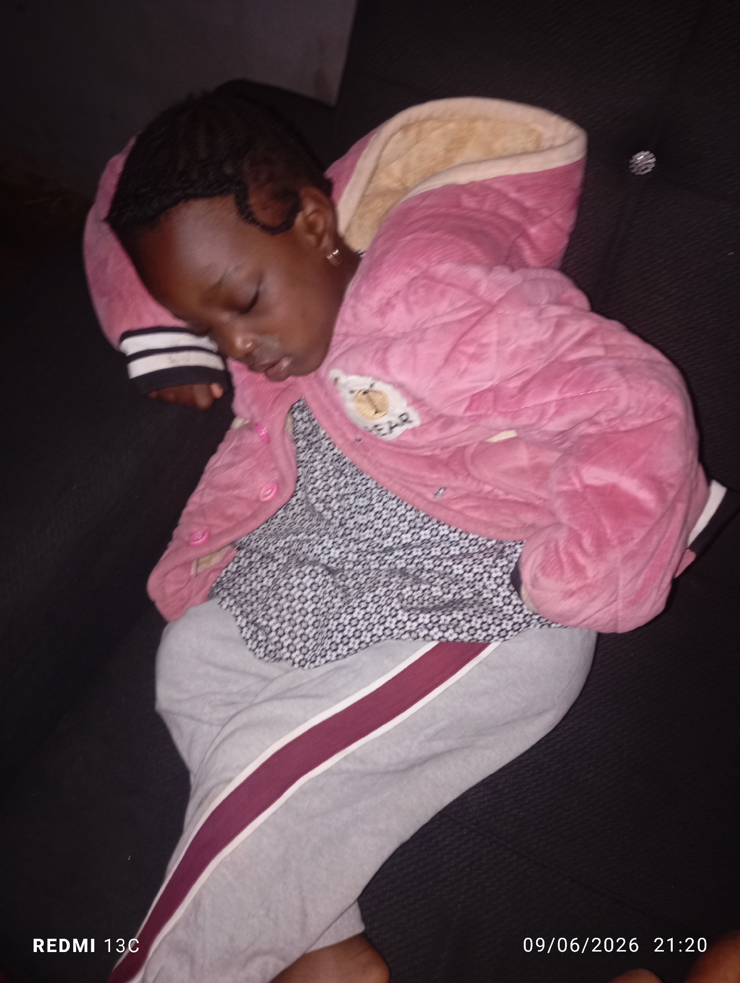 A child is sleeping on a dark-colored couch. The child is wearing a pink quilted jacket with a faux fur lining, a grey patterned shirt, and grey sweatpants with a maroon and white stripe down the side. The child's hair is braided. The image has a timestamp in the bottom right corner that reads "09/06/2026 21:20" and a device name in the bottom left corner that reads "REDMI 13C".