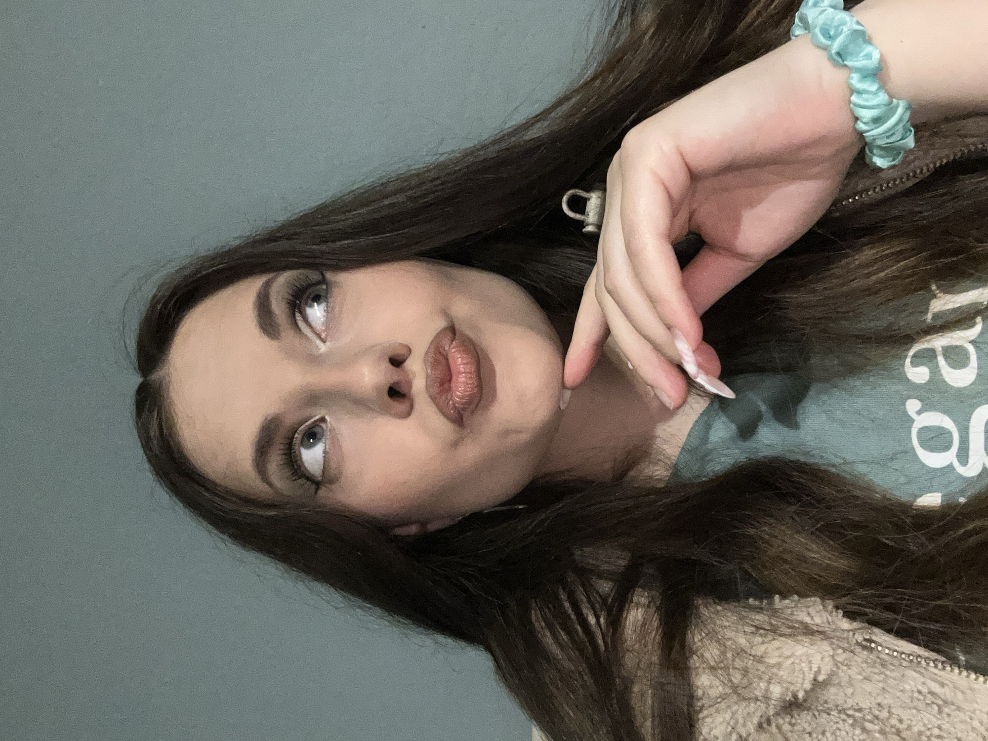 A young woman with long, dark hair is posing for a photo. Her face is tilted upward and she is looking off-camera. She is wearing makeup, including false eyelashes and lip gloss. Her hand is raised to her face. She is wearing a light blue scrunchie on her wrist. She is wearing a blue t-shirt and a tan jacket. The background is a light gray wall. The location is Unknown, Unknown.