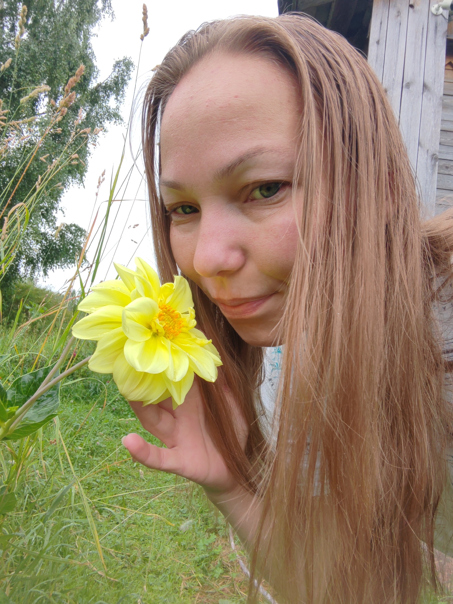 A young woman with long, light brown hair is in a grassy outdoor setting, holding a bright yellow flower to her nose. She is looking at the camera with a slight smile. Her right hand is supporting the flower, and her left hand is not visible. Behind her, there is a wooden structure to the right and green trees and tall grass to the left. The overall impression is one of a casual moment in a rural or suburban environment. The location is identified as Chukhlem, Russia, but no specific landmarks or identifying features of the city are visible in the image.