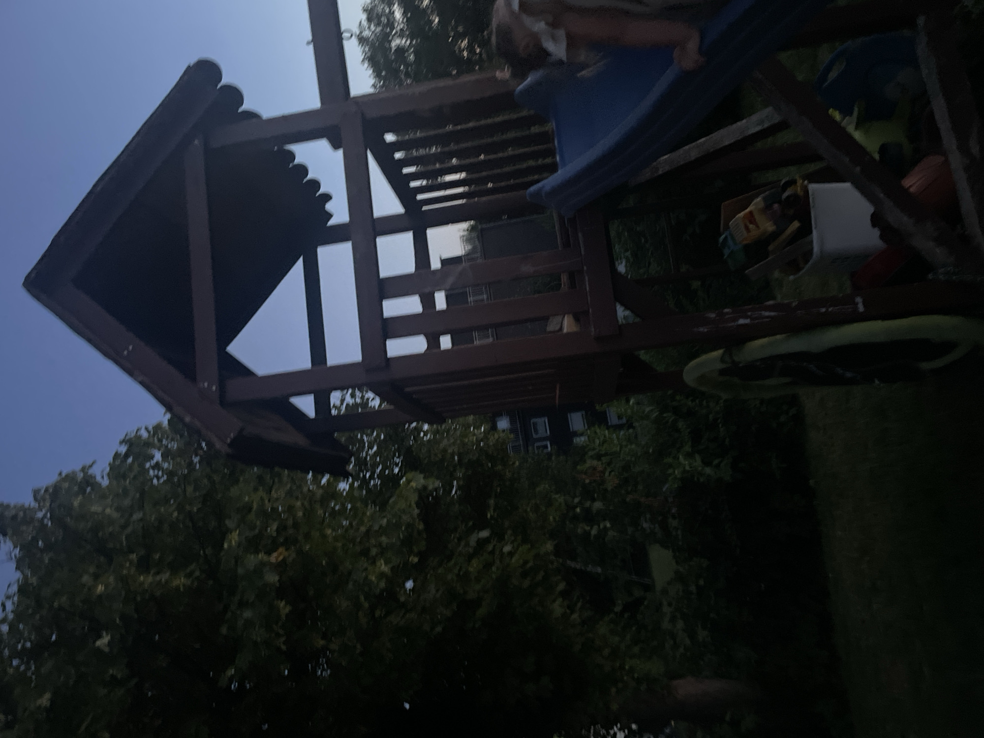 The image shows a wooden playground structure with a blue slide in the backyard of a residential building. The structure is brown and has a pointed roof. There are some toys in a white bin under the slide. A child is partially visible on the slide. The background shows a large tree and a building with windows. The sky is dark blue, indicating it is evening or dusk. The location is the city of Nümbrecht, Germany.