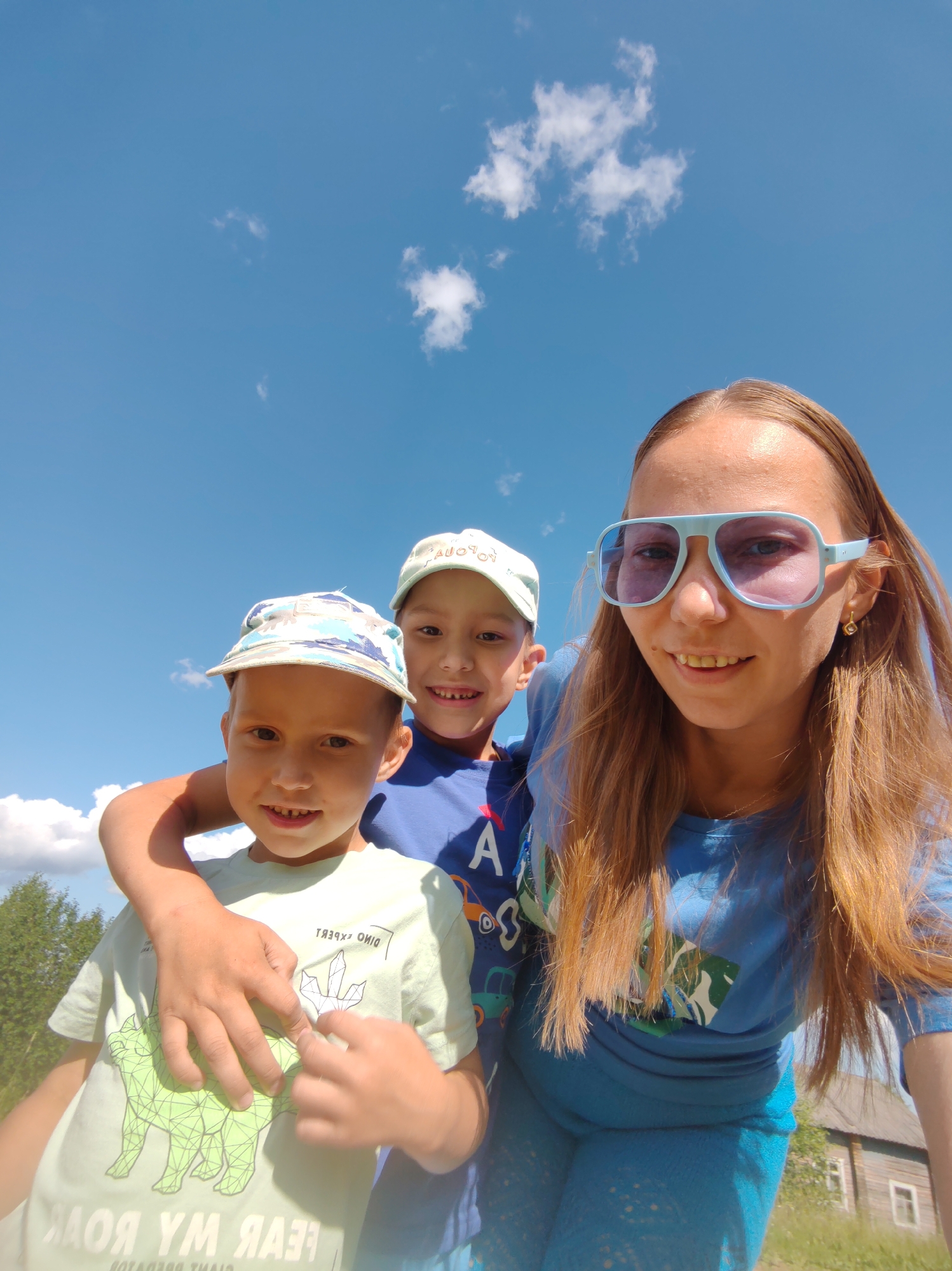 In an outdoor, potentially rural setting under a clear blue sky with scattered white clouds, a woman with long, auburn hair and light blue sunglasses is taking a selfie. She is wearing a blue t-shirt and blue patterned leggings. To her left and slightly behind her, two boys are visible. The boy closest to the camera, on the left, is wearing a light green t-shirt with a green dinosaur graphic and text, and a camouflage-patterned cap. He has his arm around the other boy. The second boy, slightly further back and to the right, is wearing a blue t-shirt and a white cap. Both boys are smiling. The background includes green trees and a wooden building. The image appears to be taken in daylight.